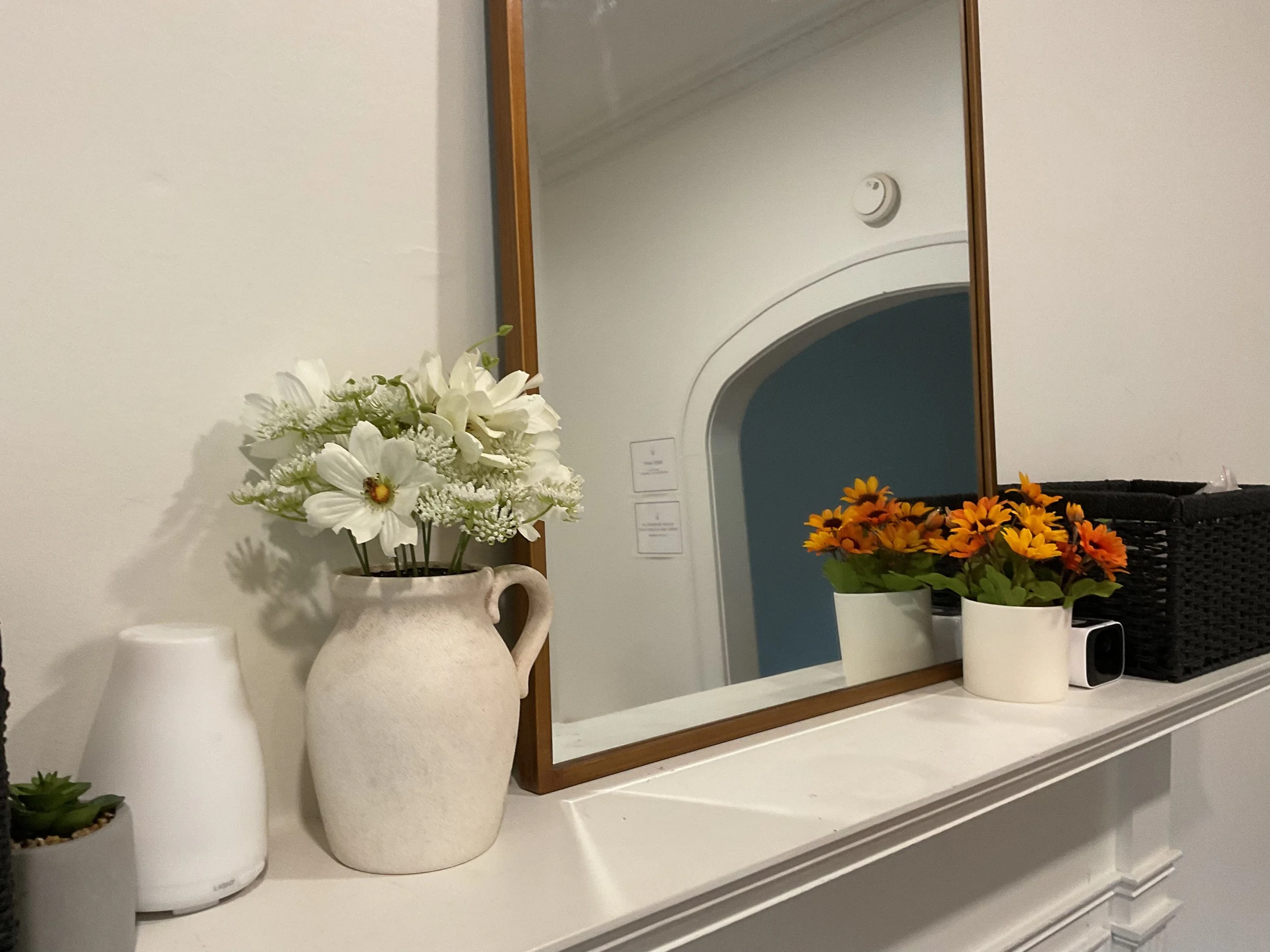 Decorative white ceramic vases with artificial flowers on a white mantelpiece, with a large mirror reflecting a white wall with a smoke detector and a doorway.