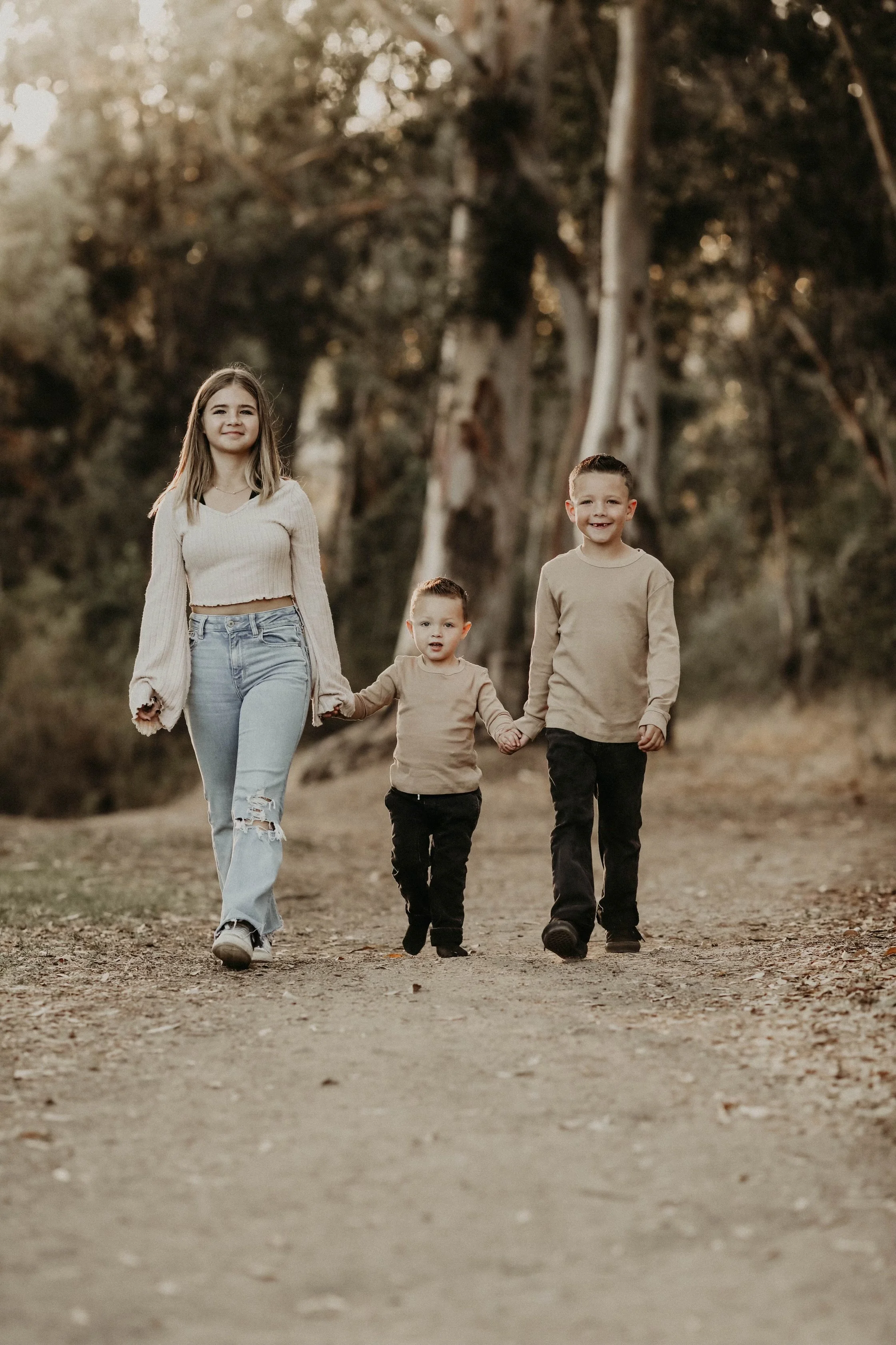 A girl and two boys walking hand in hand on a dirt path in a forest during sunset.