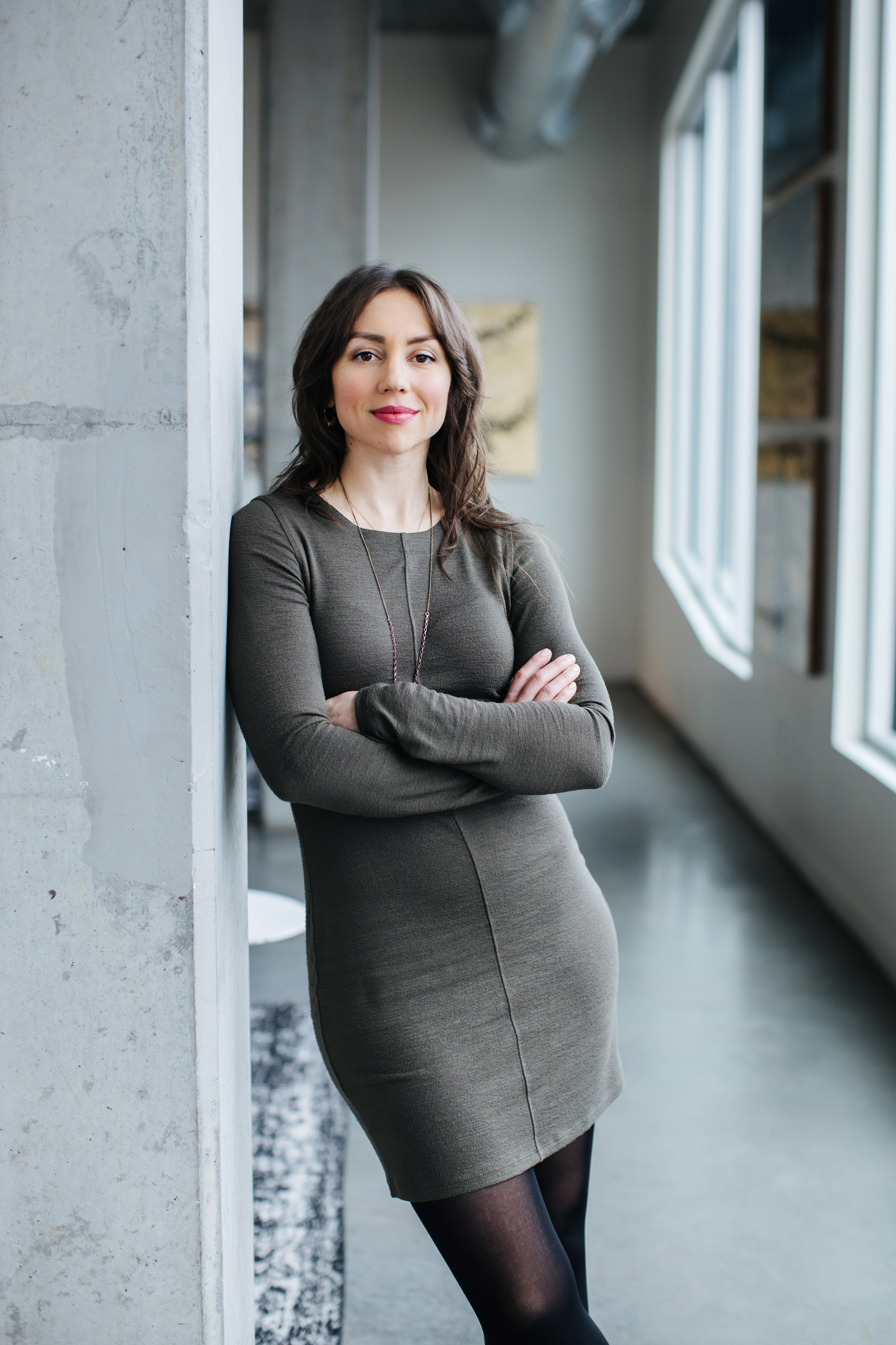 A female multidisciplinary researcher and DJ with dark hair, wearing a grey dress and black tights, leaning against a concrete wall with arms crossed, standing in a modern, well-lit space with windows.
