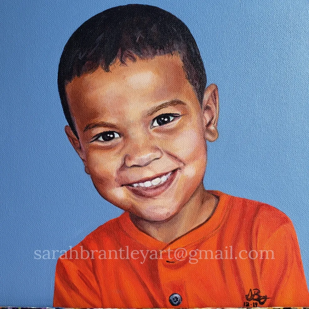 A portrait of a young boy with short hair, smiling, wearing an orange shirt against a blue background.