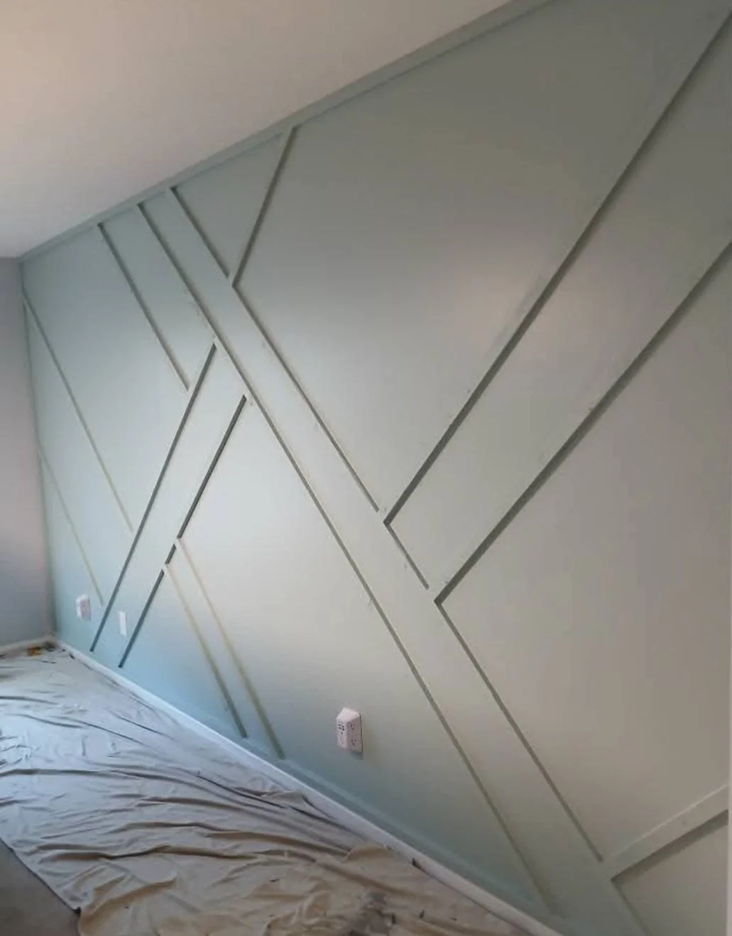 Interior wall with geometric paneling in a light green color, with electrical outlets and a protective sheet on the floor.