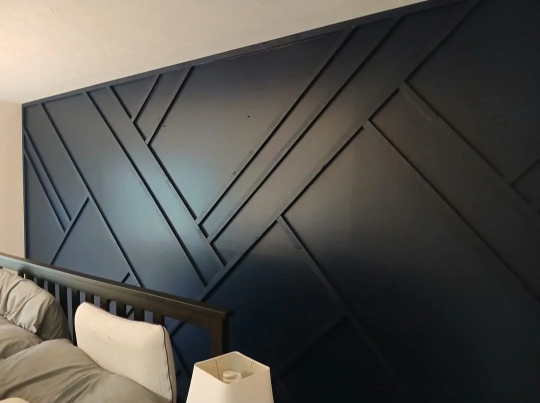Close-up of a bed with a beige headboard, beige bedding, a beige pillow, and a black headboard wall with geometric paneling.