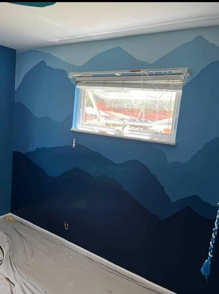 Interior of a room with mountain silhouette mural painted on the blue wall, window with blinds, and painting supplies on the floor.