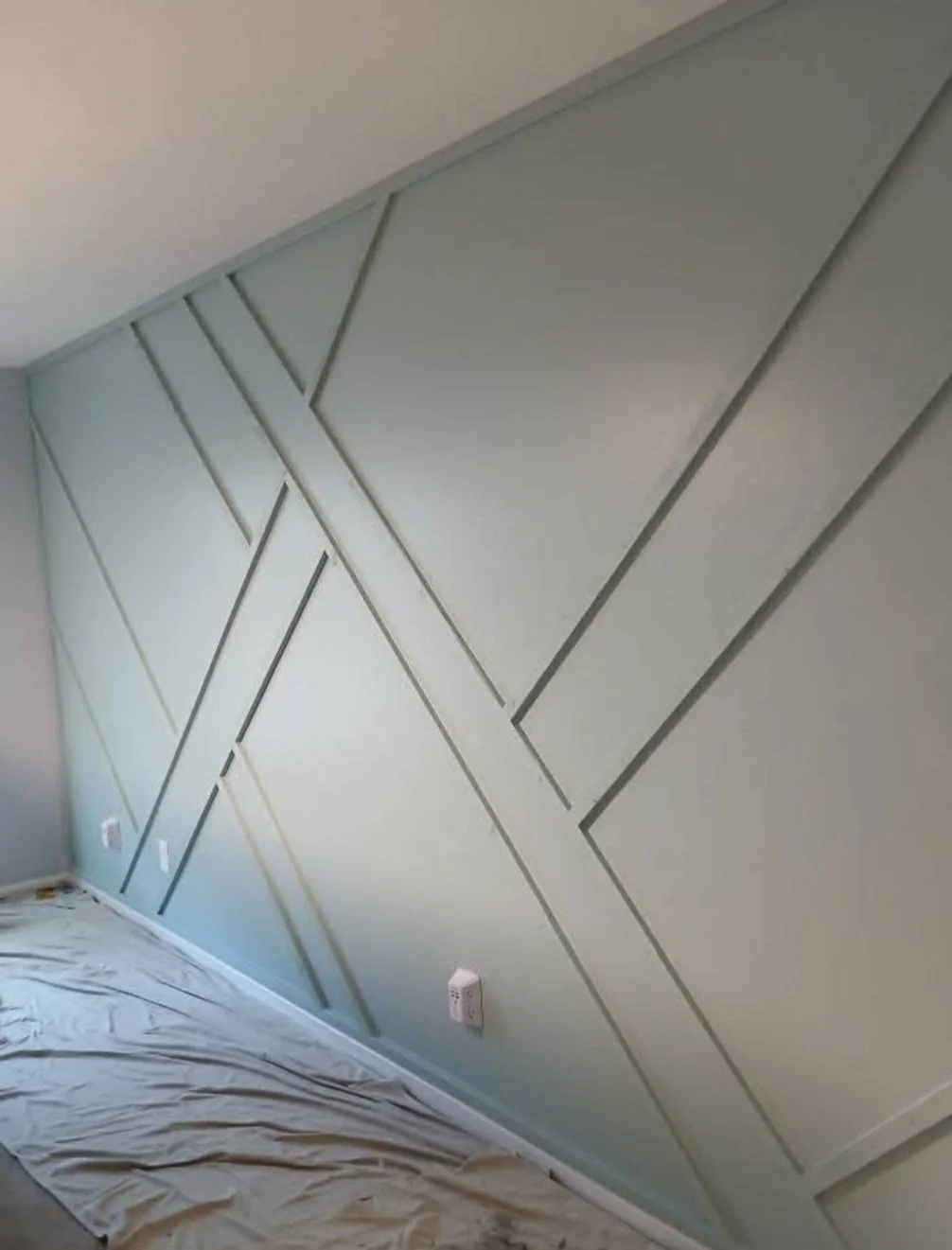 Interior wall with geometric paneling design, painted light green, with electrical outlets and a plastic sheet on the floor.