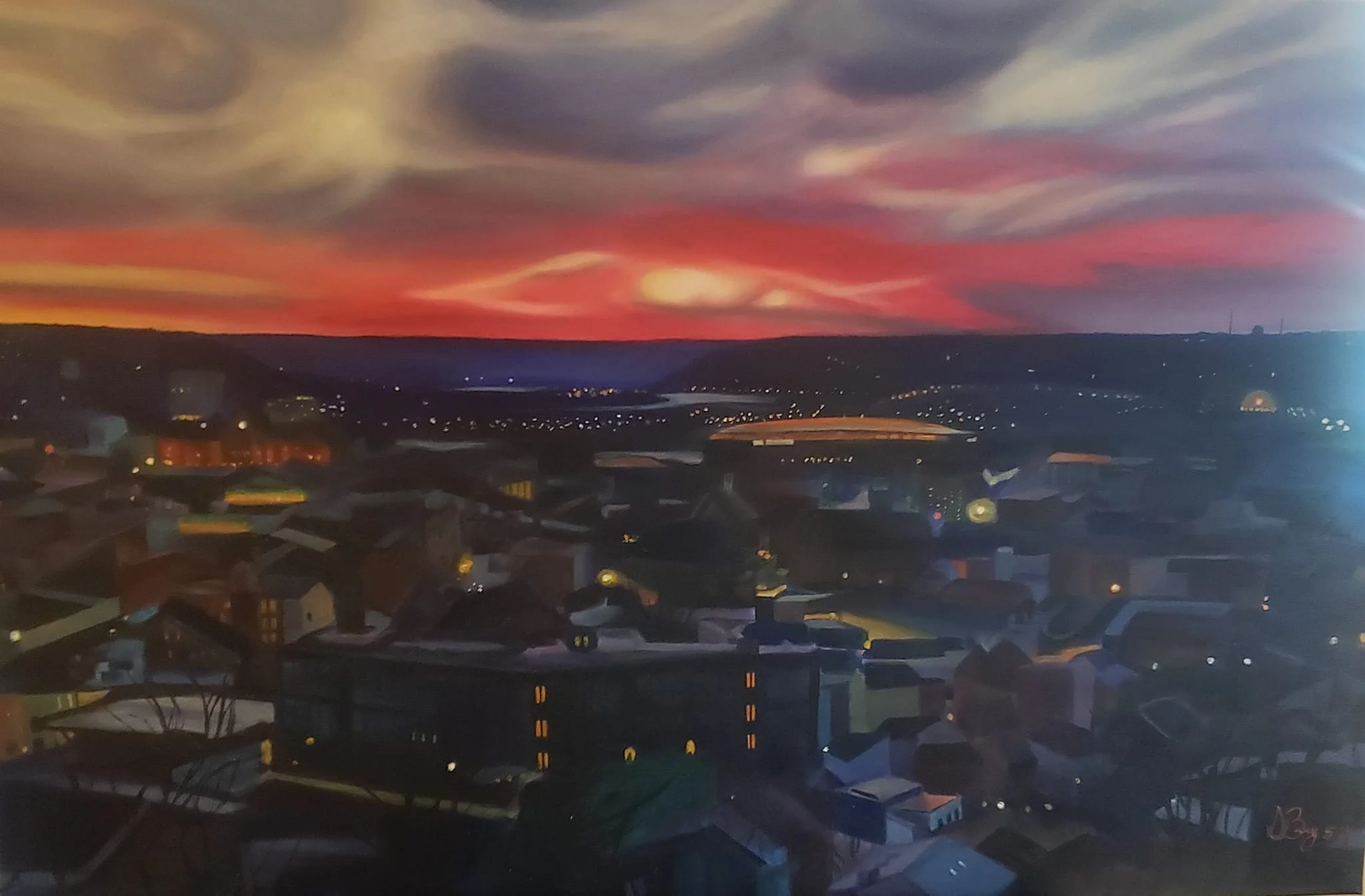 Colorful sunset over a cityscape with houses and buildings in the foreground, and distant hills or mountains under a sky with swirling clouds.