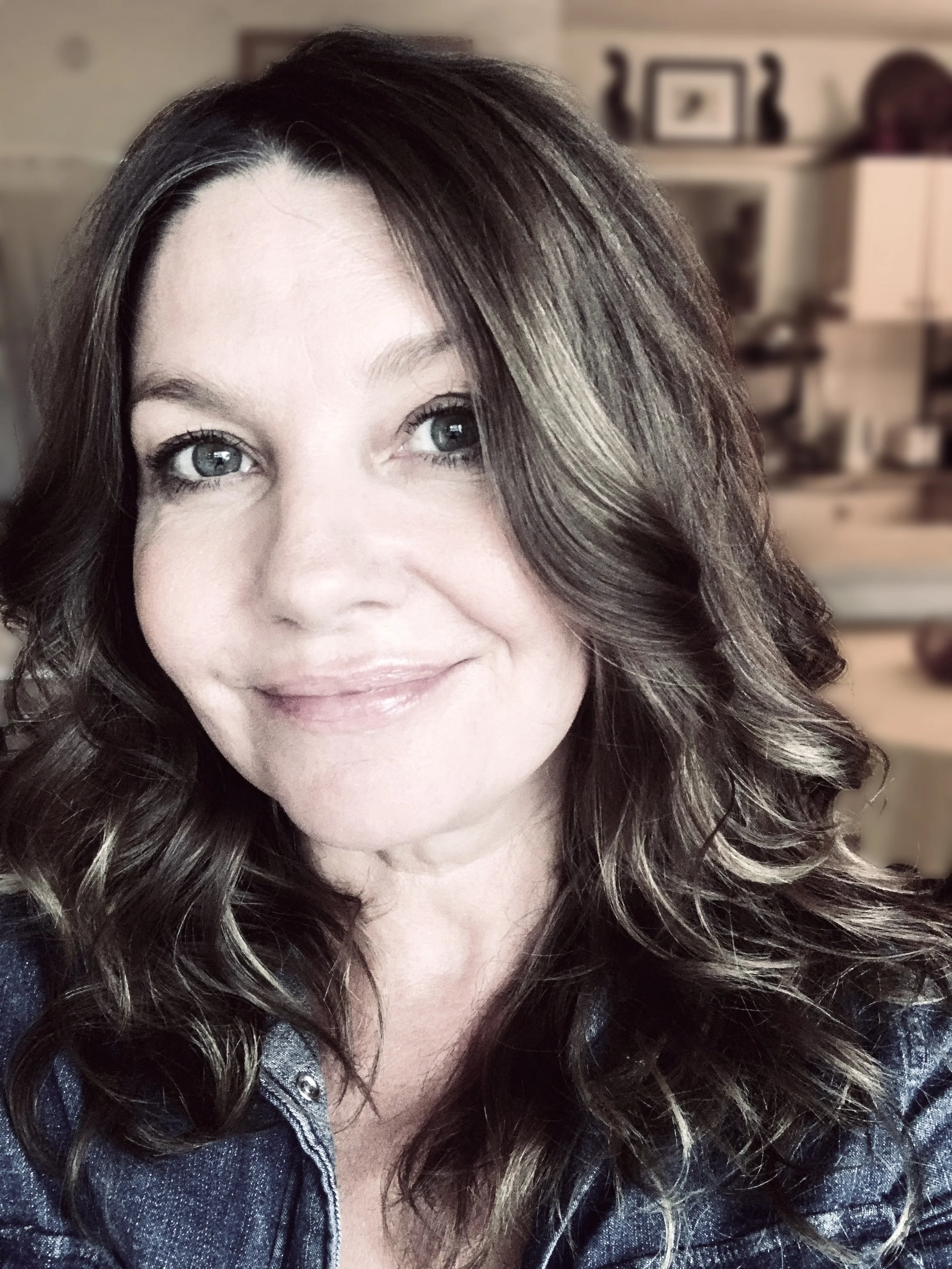 A female coach in Thirroul, Australia, smiles warmly. Close-up of a woman with light-colored eyes and wavy brown hair, wearing a blue denim jacket, smiling softly, indoors with blurred background.