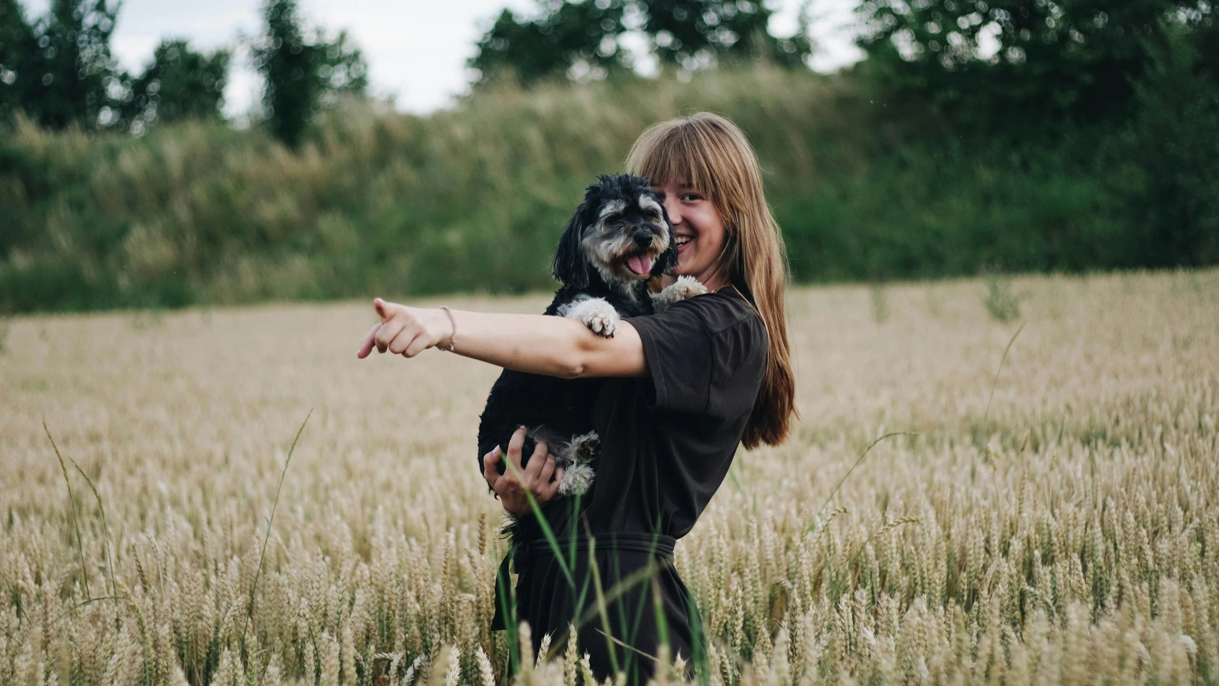 A young woman with long red hair smiling and holding a small black and white dog in a wheat field during daytime.