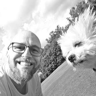 The owner of Cheery Beauties with glasses smiling next to Ollie outdoors on a paved area with trees in the background.