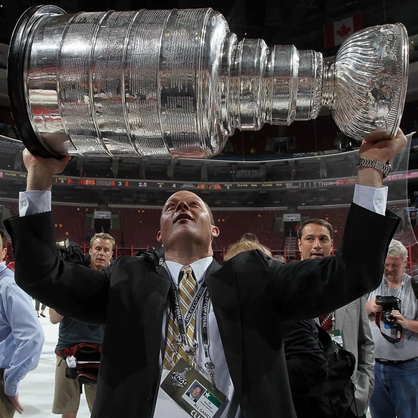 A man in formal attire holding up the Stanley Cup trophy after winning a hockey championship, with several other people and a hockey arena in the background.