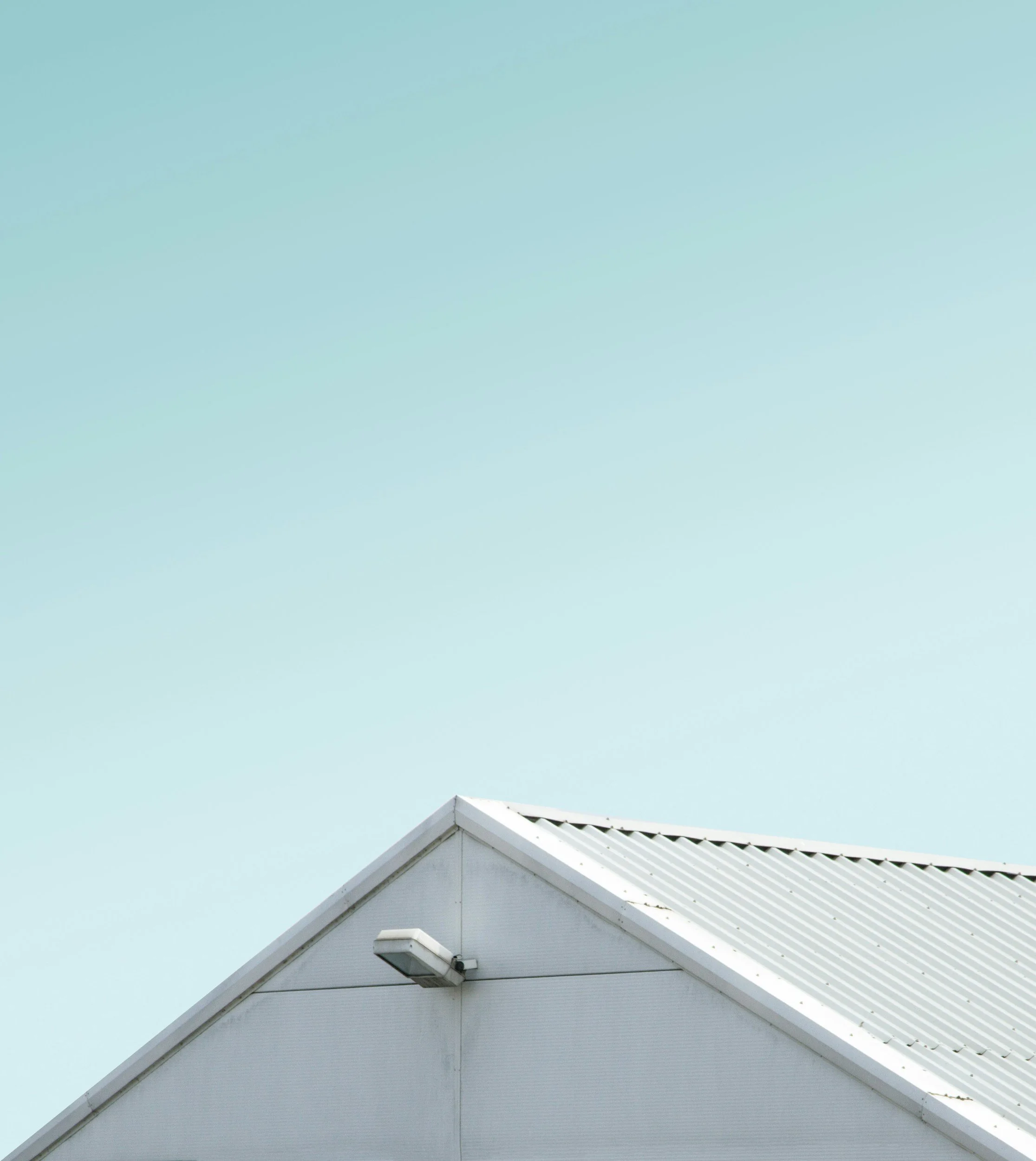 A portion of a white house roof with a gable end, a white gutter, and a security light, set against a light blue sky.