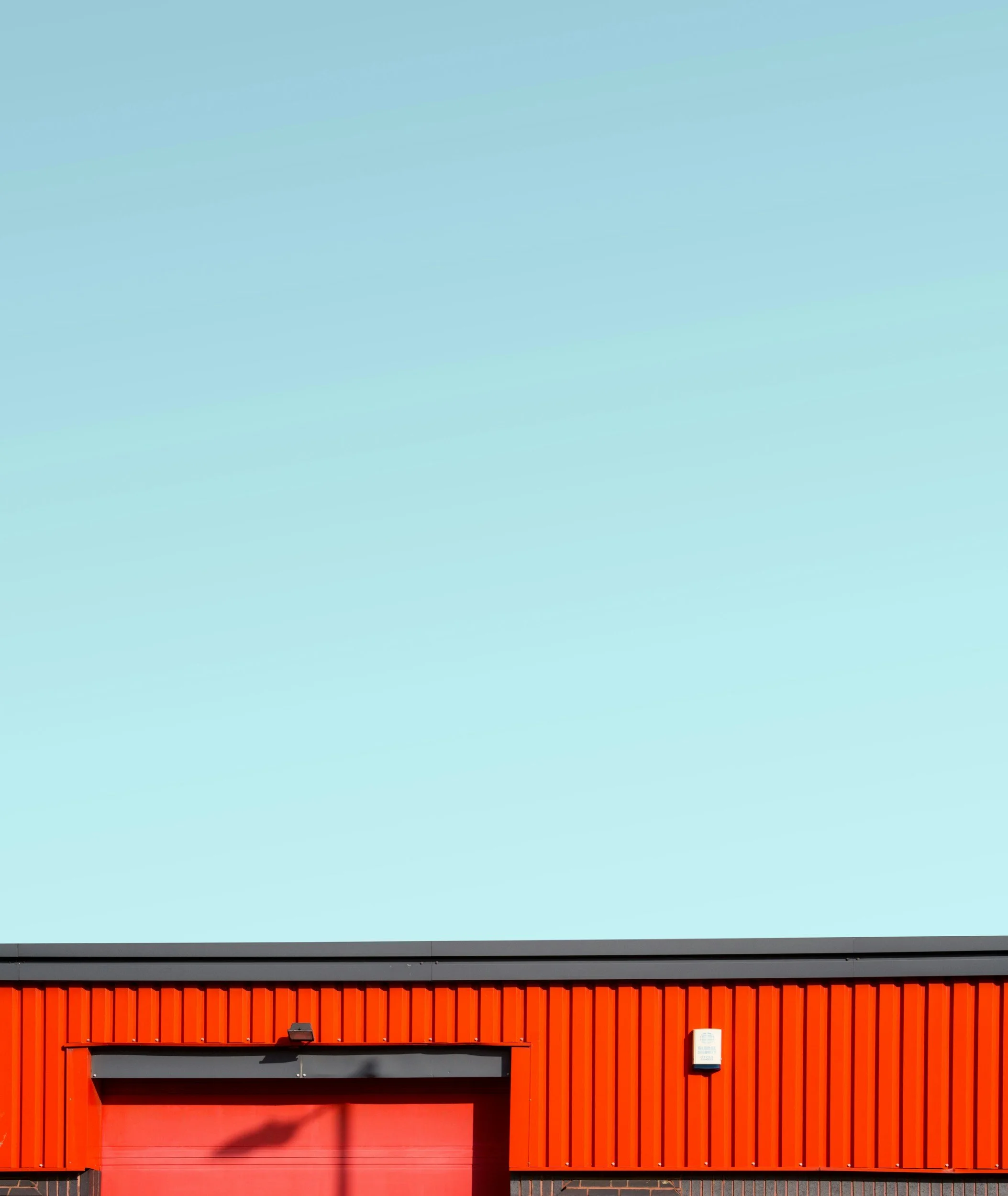 Close-up of a modern commercial building with a red corrugated metal facade, gray trim, and a closed red garage door. There is a small sign and security camera mounted on the wall.