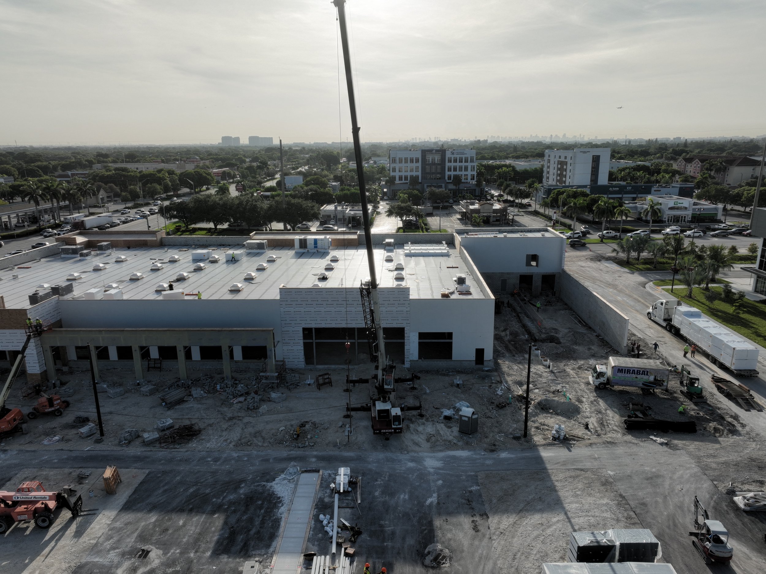 Construction site with workers, heavy machinery, and building materials, in an urban area with trees and buildings in the background.