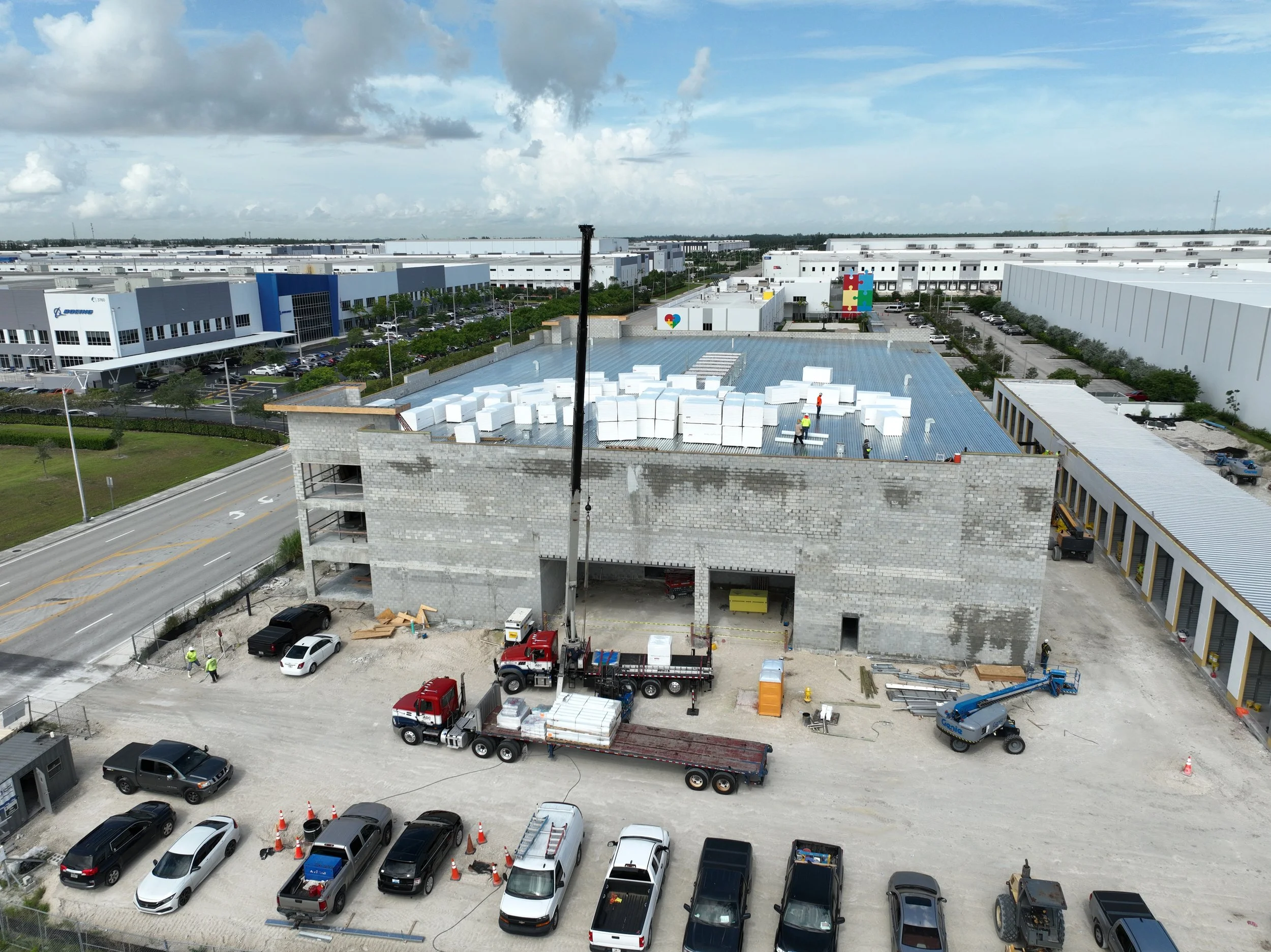 Construction site with workers and trucks, surrounded by commercial buildings under a partly cloudy sky.