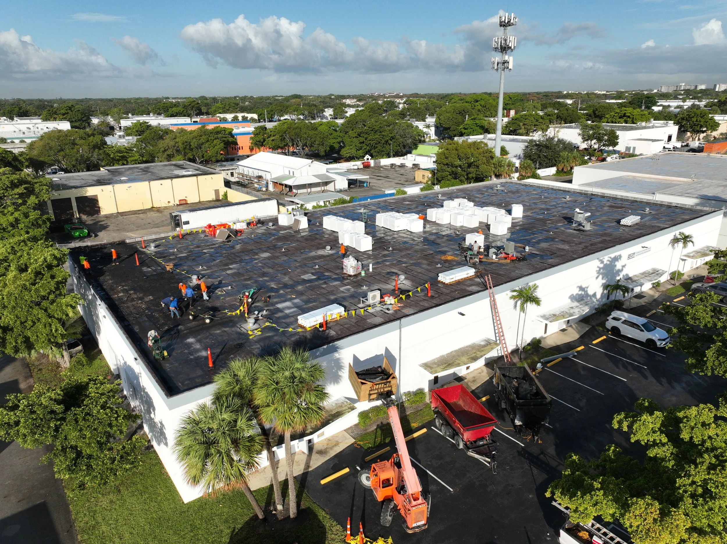 An aerial view of a commercial building's flat roof undergoing maintenance or repair, with workers, orange cones, equipment, and a crane, surrounded by trees and nearby buildings under a partly cloudy sky.