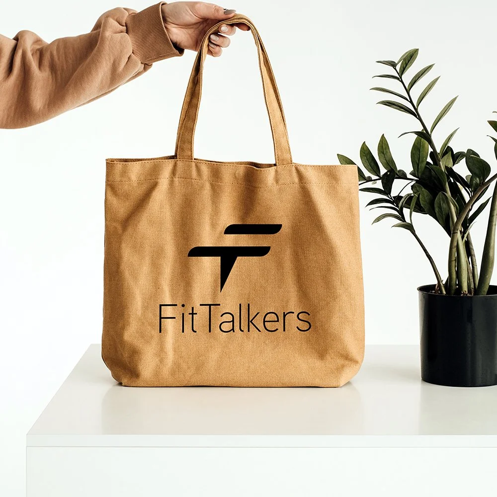 A tan tote bag with the FitTalkers logo, featuring a speech bubble icon, sitting on a white table next to a potted plant.