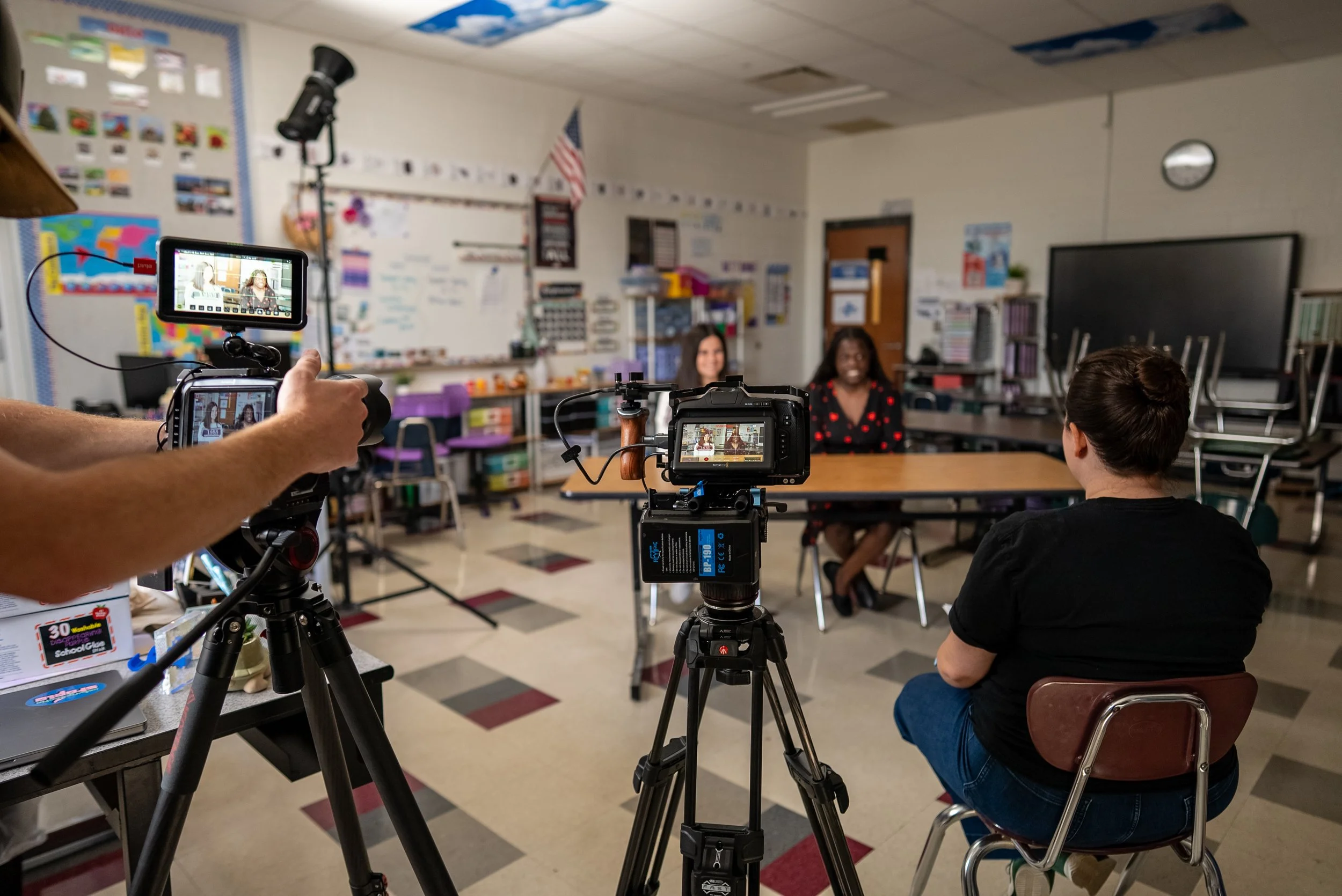 A classroom with two women being recorded for a video or interview, with cameras focused on them. One woman is seated at a desk, and another is sitting nearby. Classroom decorations and furniture are visible in the background.