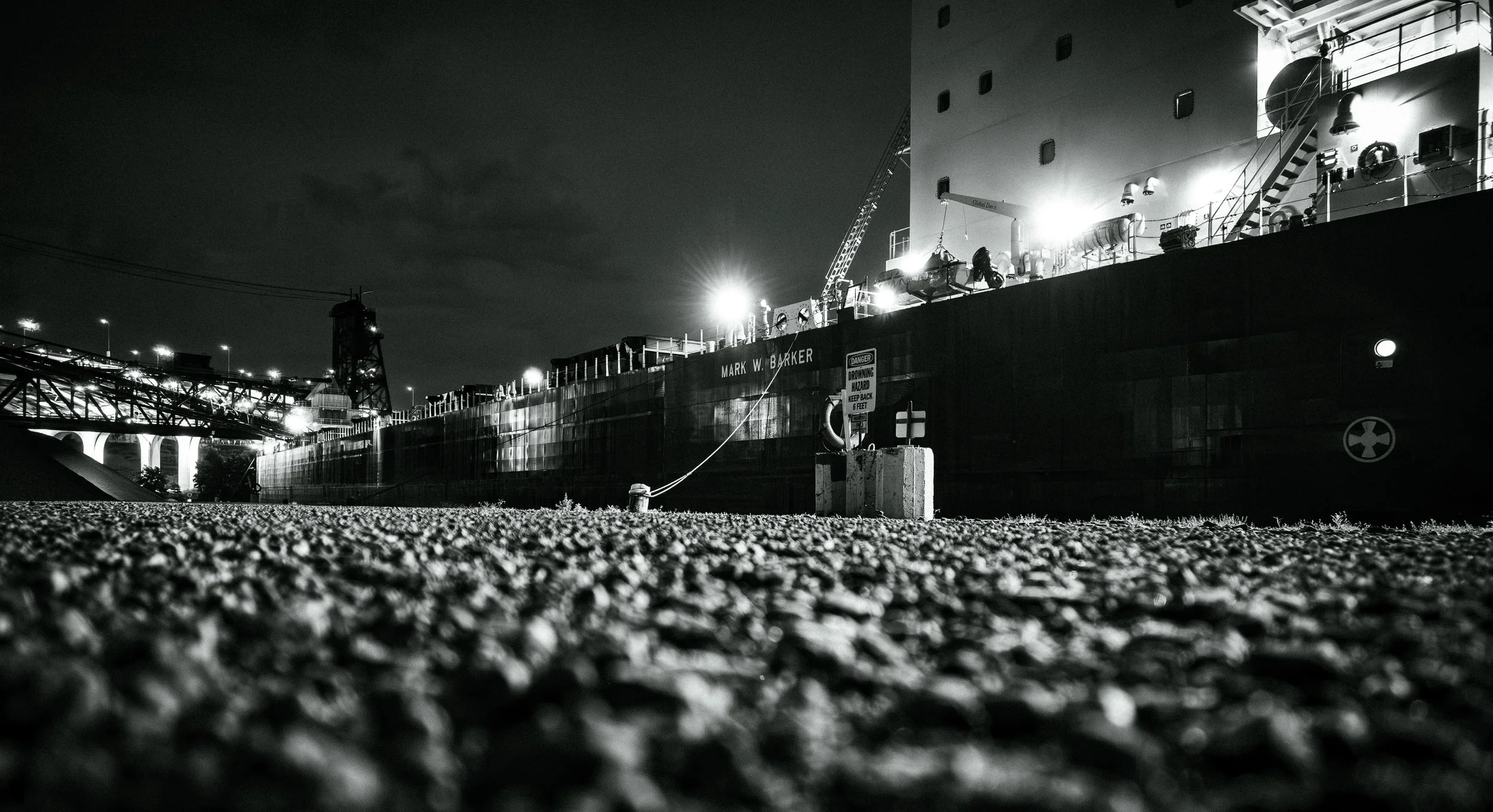 A black-and-white photograph of a large ship docked at a port, illuminated by bright lights, with a gravel ground in the foreground.
