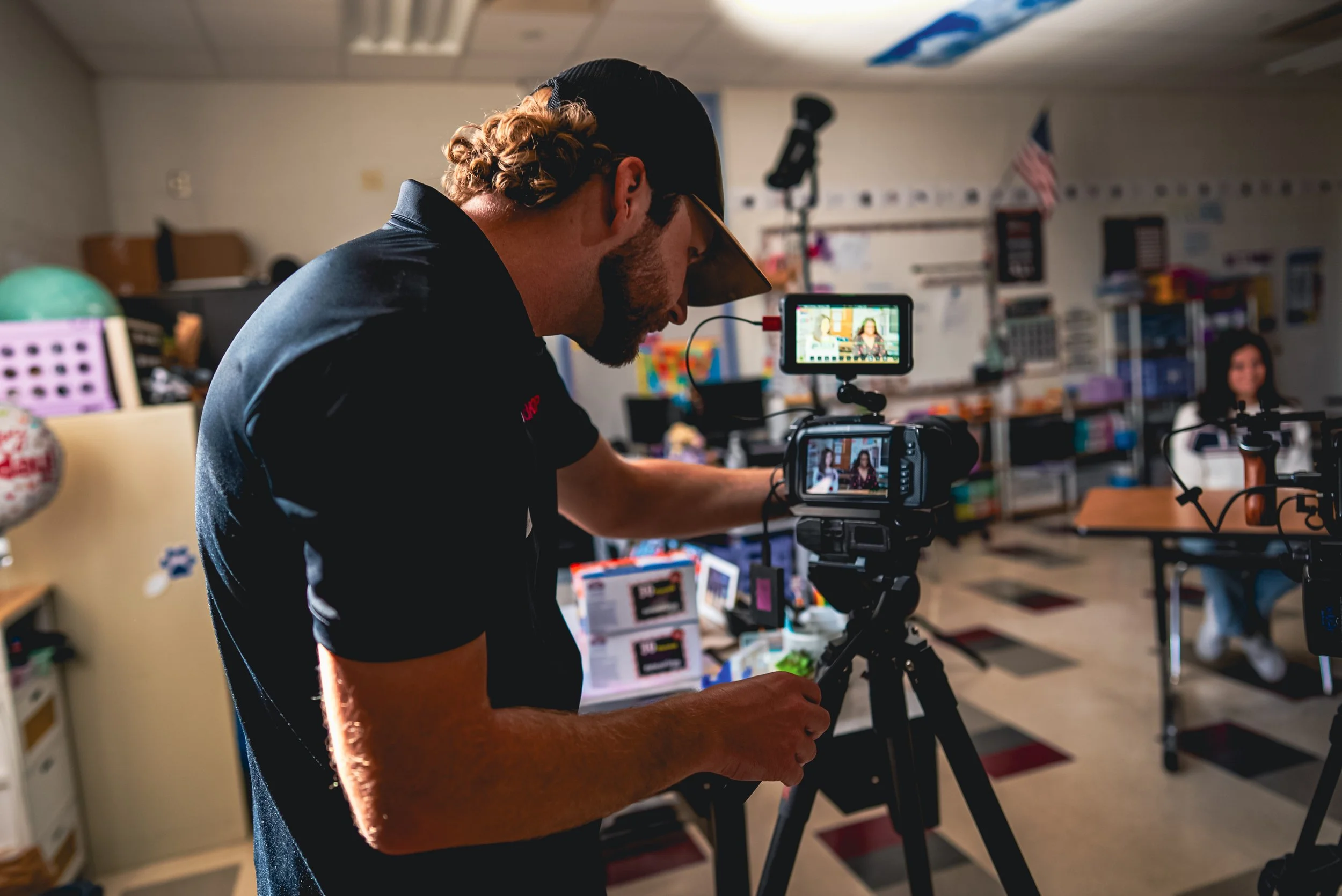 A man operating a professional video camera in a classroom or studio environment, with a woman sitting in the background, surrounded by classroom supplies and equipment.
