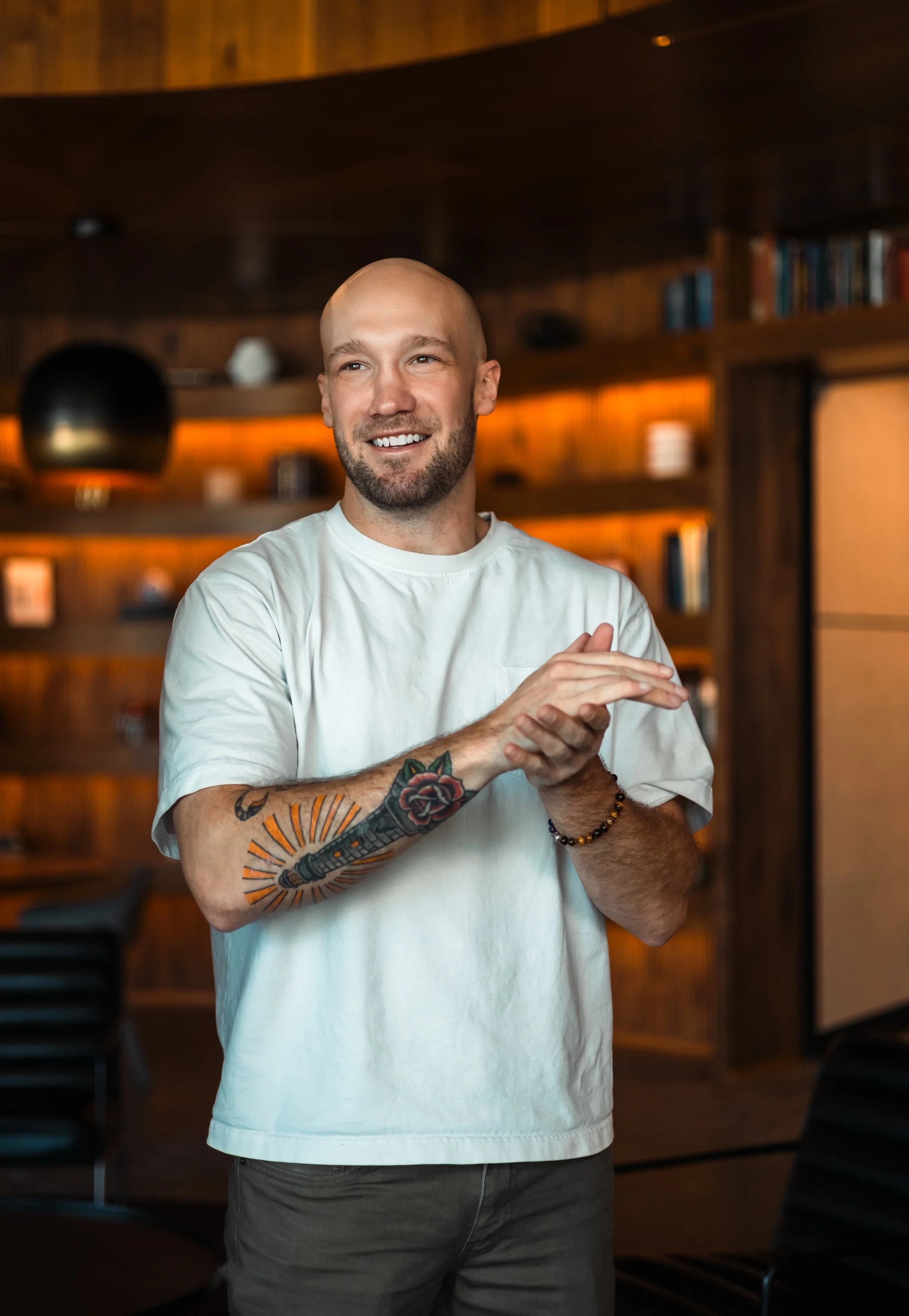 A smiling bald man with tattoos on his left arm, wearing a white t-shirt and dark pants, standing in a warmly lit room with wooden shelves and books in the background.