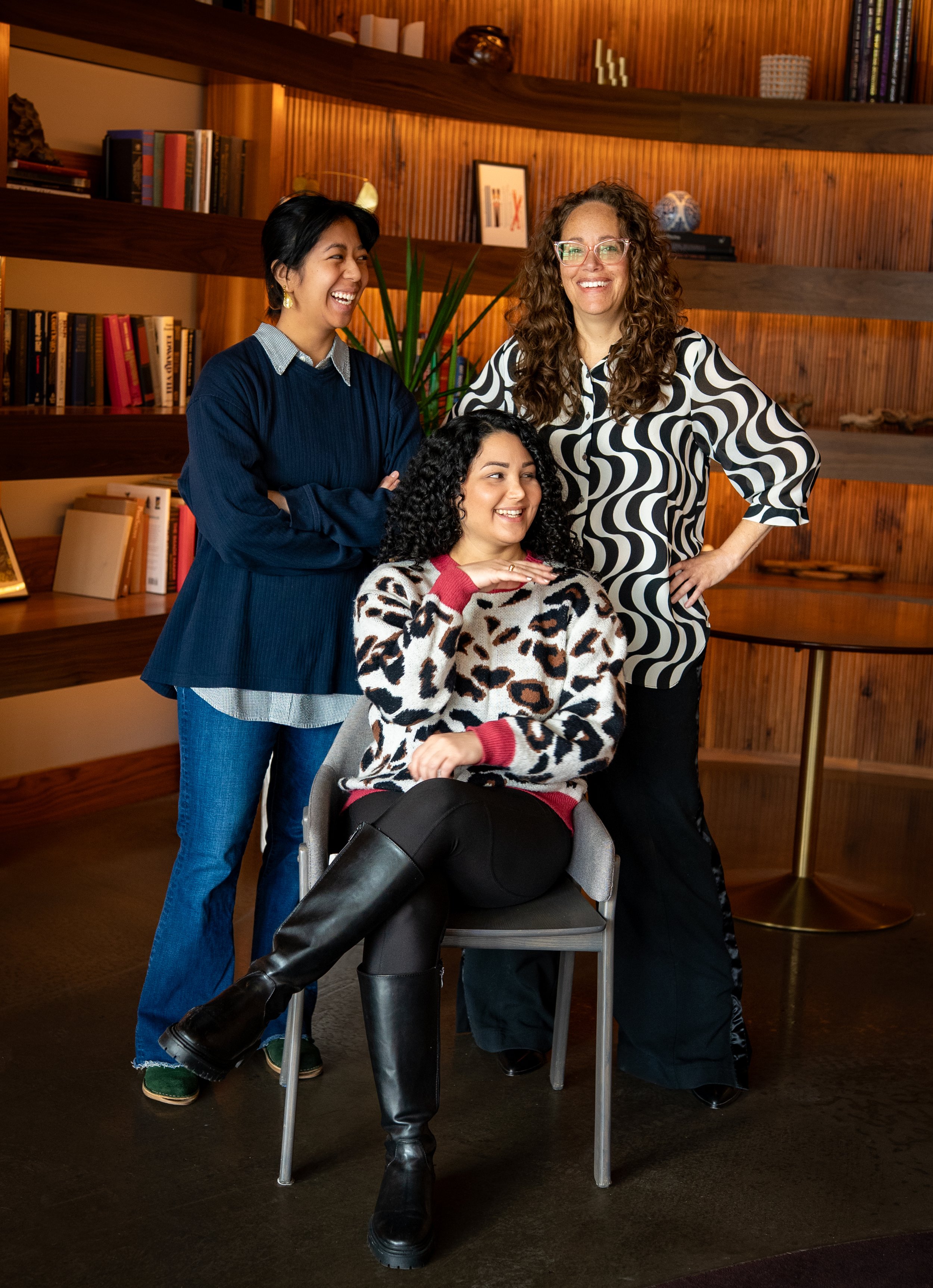 Three women smiling and posing together in a cozy room with wooden shelves and a dark wooden desk, one sitting on a chair and the other two standing behind her, dressed in stylish casual clothing.