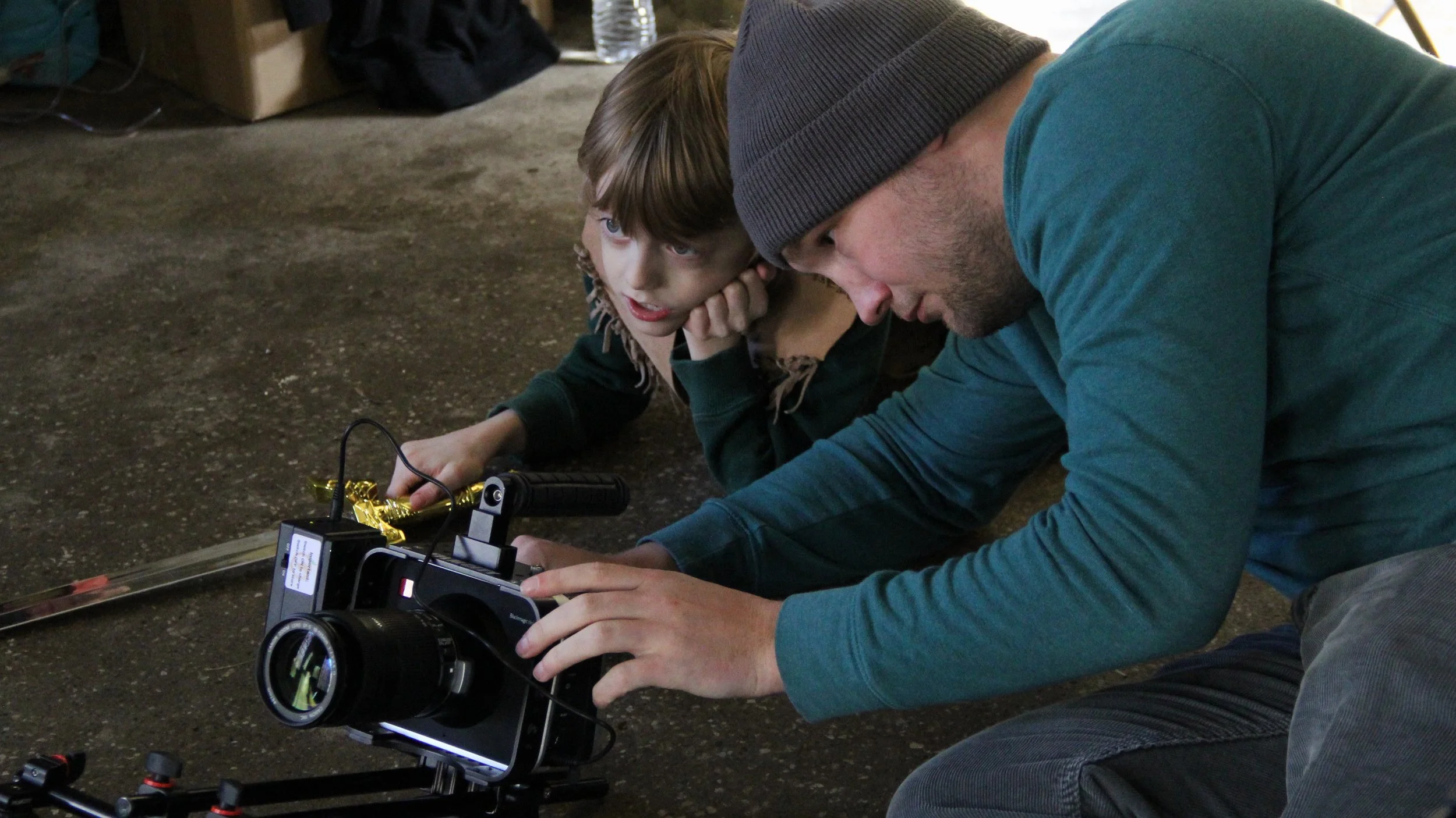 A man and a young boy lying on the ground, looking at a camera mounted on a sliding rail, with a gold-colored object nearby. The boy has brown hair and is wearing a dark shirt, while the man is wearing a beanie and a teal long-sleeve shirt.