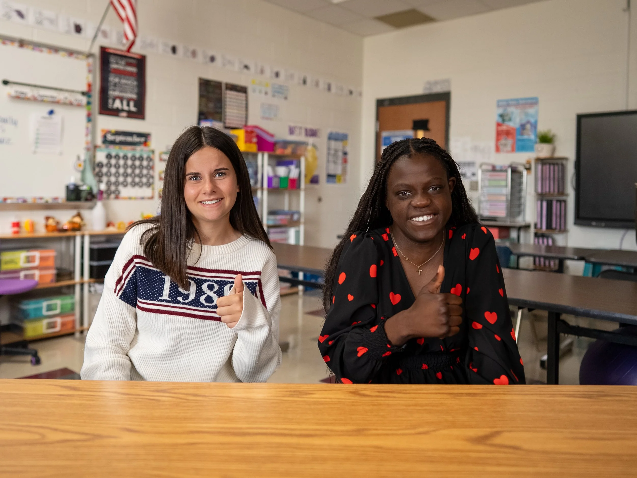 Two girls sitting at a desk in a classroom giving a thumbs-up gesture, smiling at the camera.