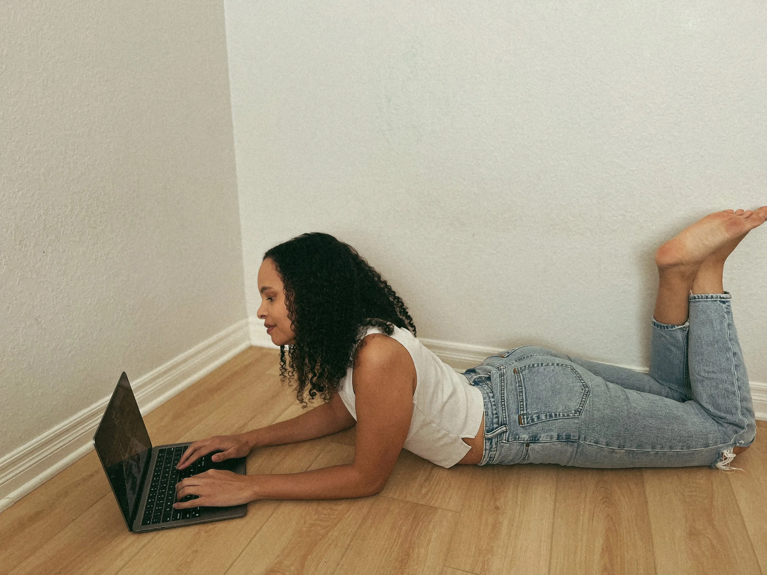 A woman with curly dark hair lying on her stomach on a hardwood floor, wearing a white tank top and blue jeans, using a laptop computer in front of a plain white wall.