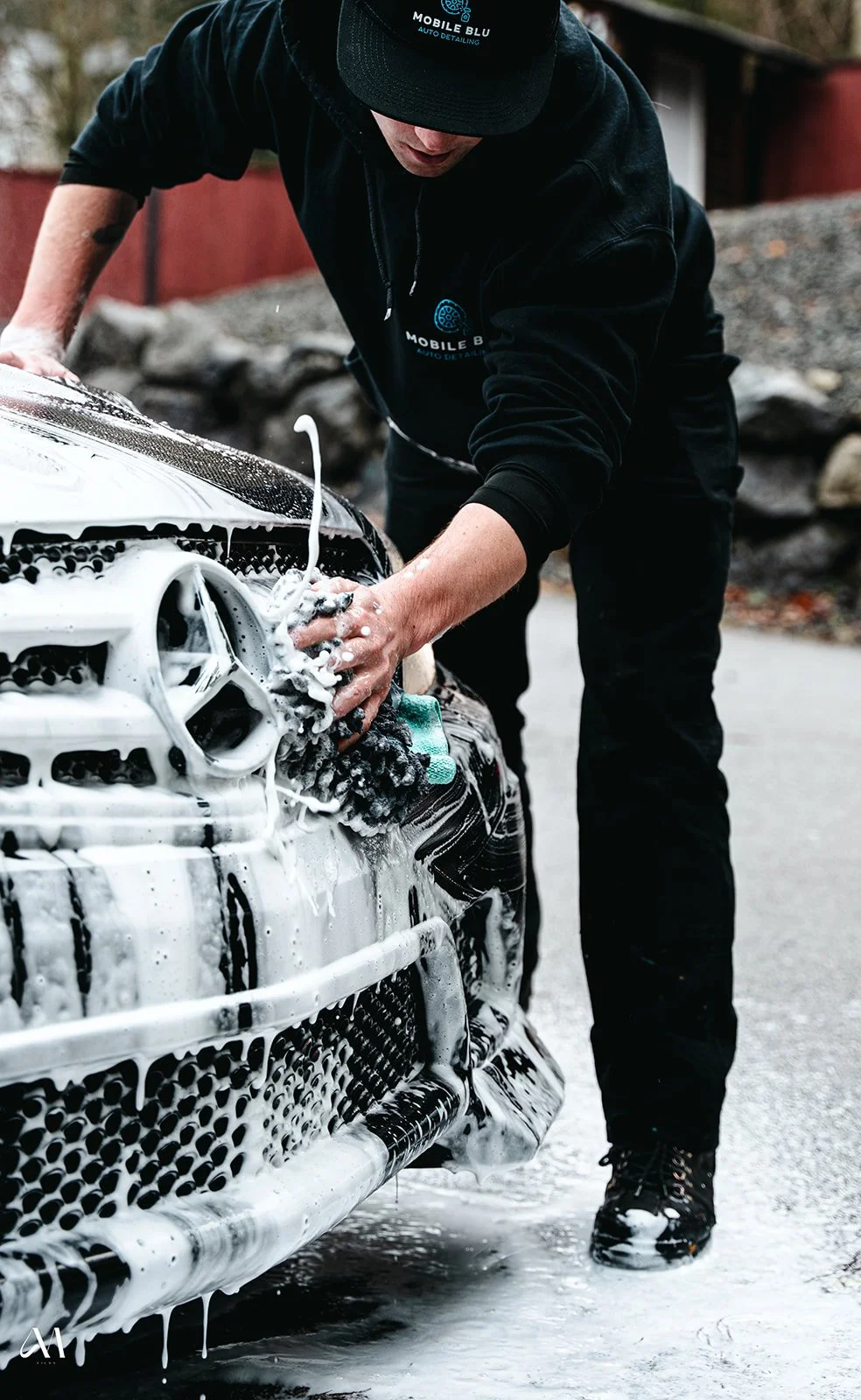 Person washing a white Mercedes-Benz car with soap and sponge, wearing a black hoodie and cap, parked outdoors with rocks in background.