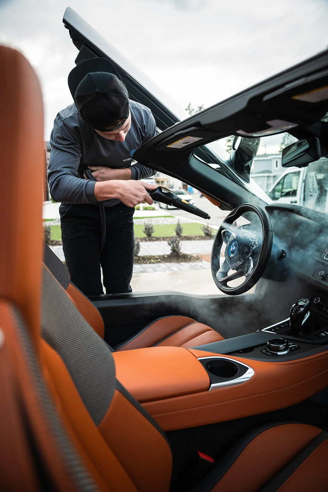 A person wearing a black cap and gray long-sleeve shirt cleaning or inspecting the inside of a car with a handheld tool, while the car's windows are steamed up, and the interior features orange and black seats.