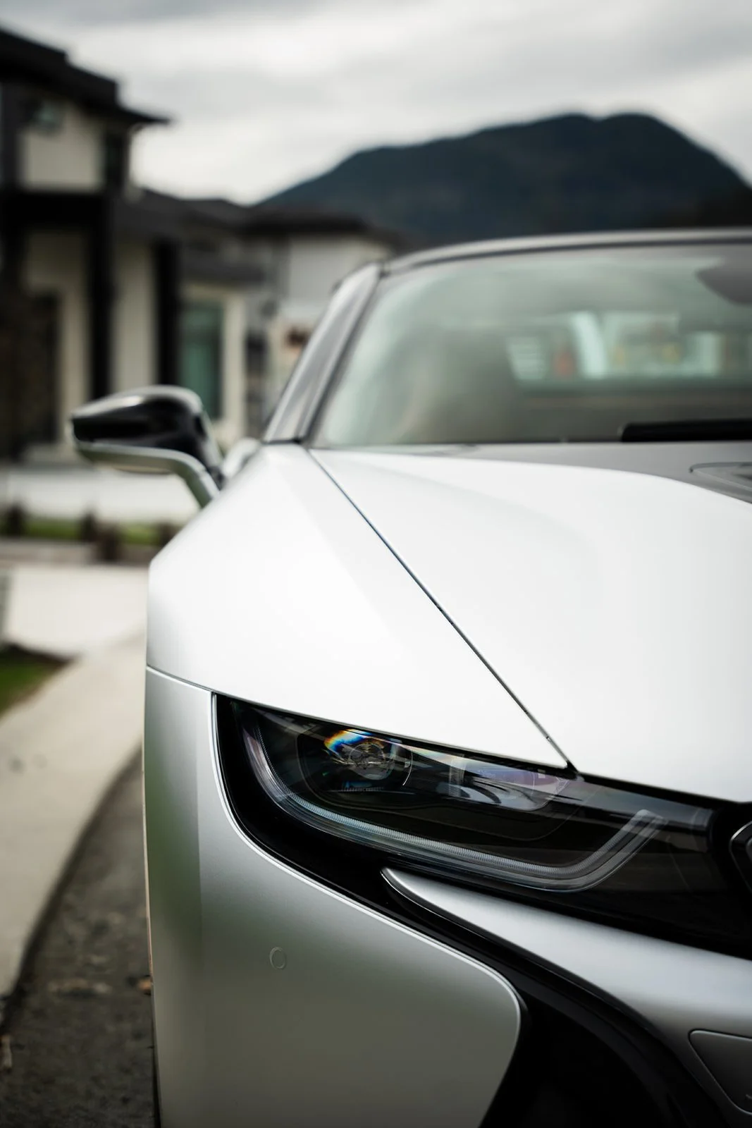 Close-up of the front left side of a silver sports car with a mountain and houses in the background.