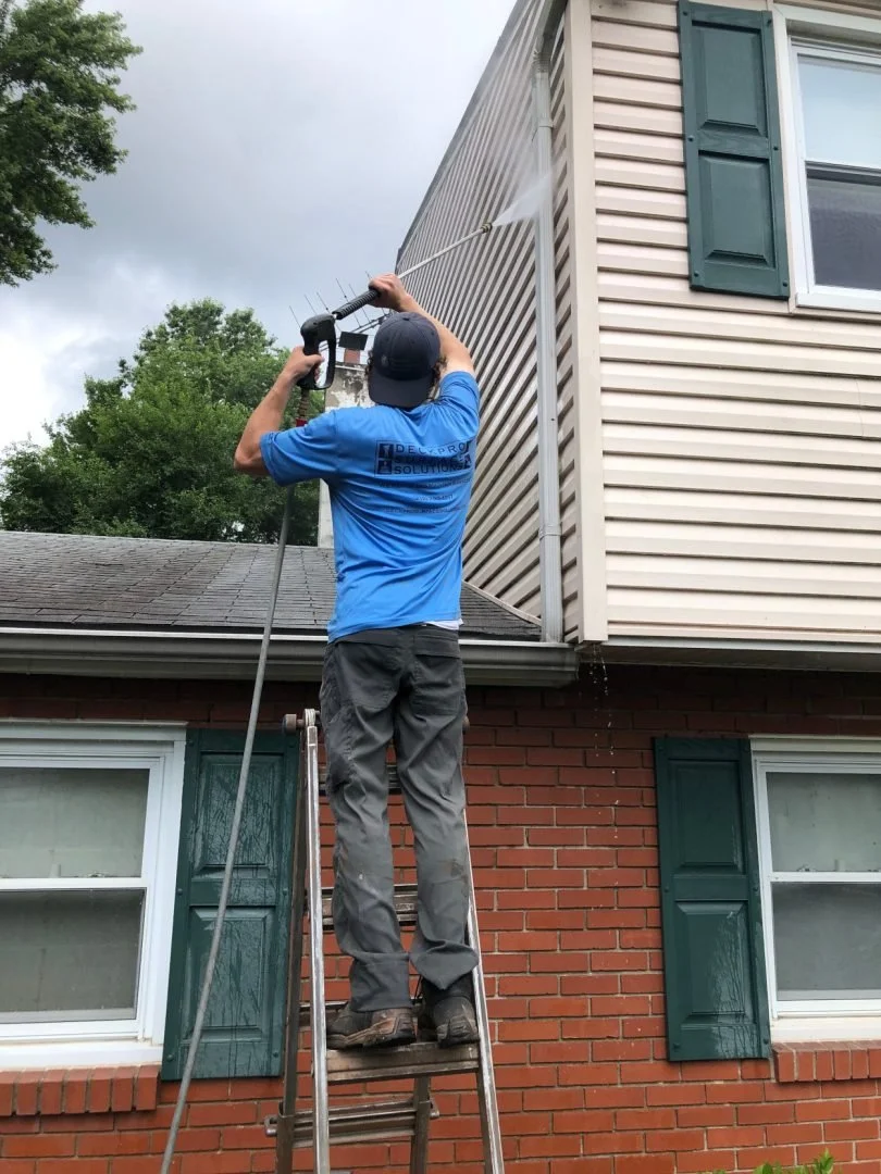 A man standing on a ladder power washing the side of a house. He is wearing a blue shirt and a black cap, and is using a pressure washer to clean the exterior of the house.