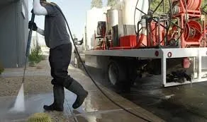 A worker in dark clothing with gloves and boots, using a hose to spray water or chemical on the ground near a truck.