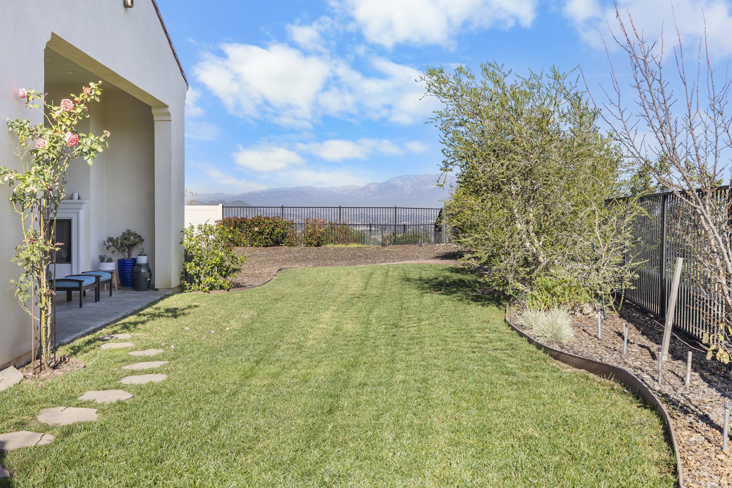 Professional real estate photography of a residential backyard with green lawn, landscaped trees, metal fencing, and mountain views designed for property listings and real estate marketing.