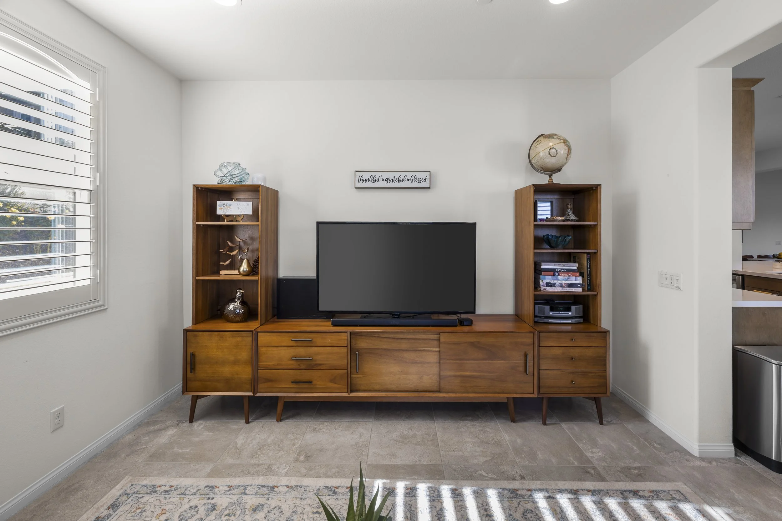 Professional real estate interior photography of a staged living room with wooden media console, shelving, flat-screen TV, and natural light for residential property marketing.