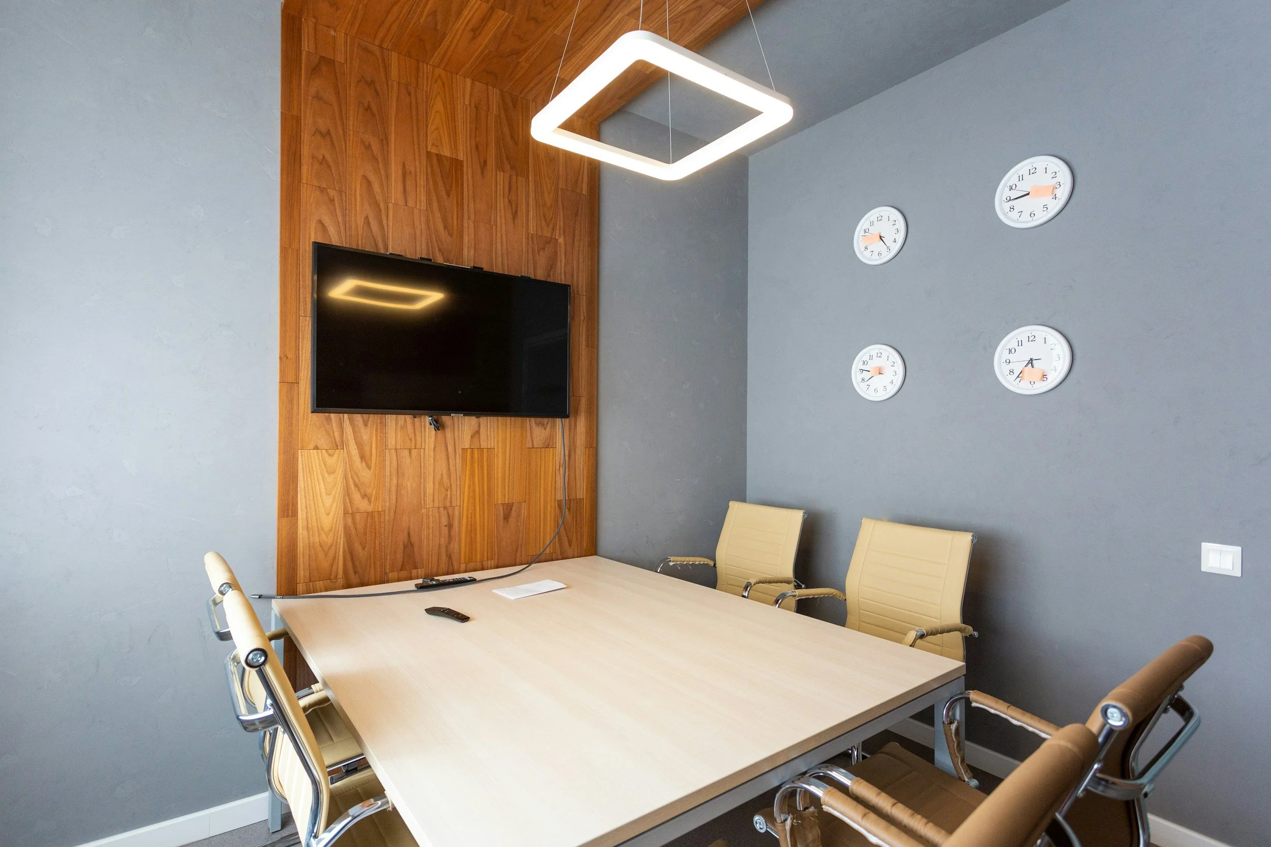 Meeting room with a light wood conference table, beige chairs, a wall-mounted TV, and wall clocks showing different times.