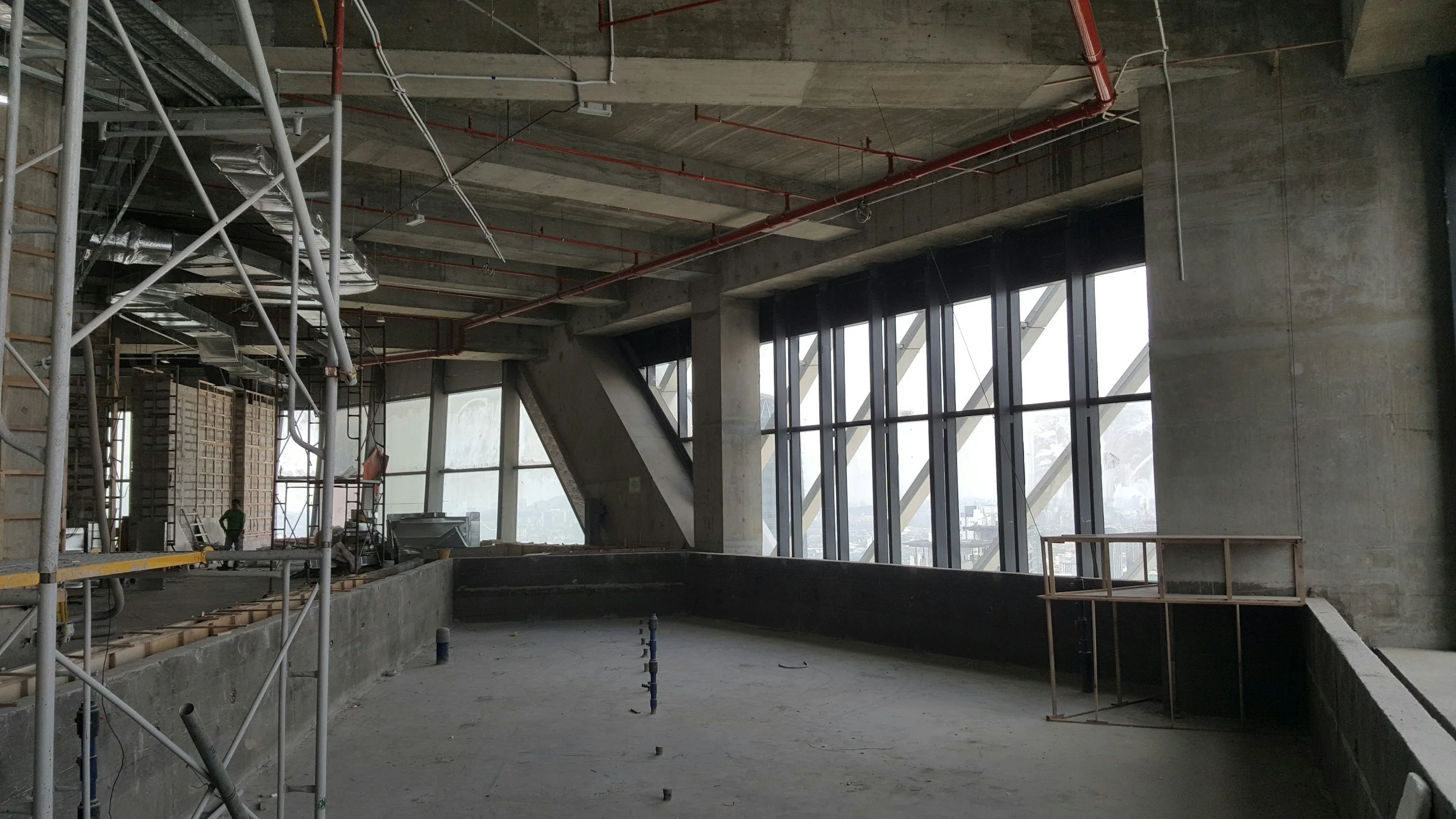 Interior view of a high-rise building under construction, showing concrete walls, large windows, scaffolding, and exposed pipes.