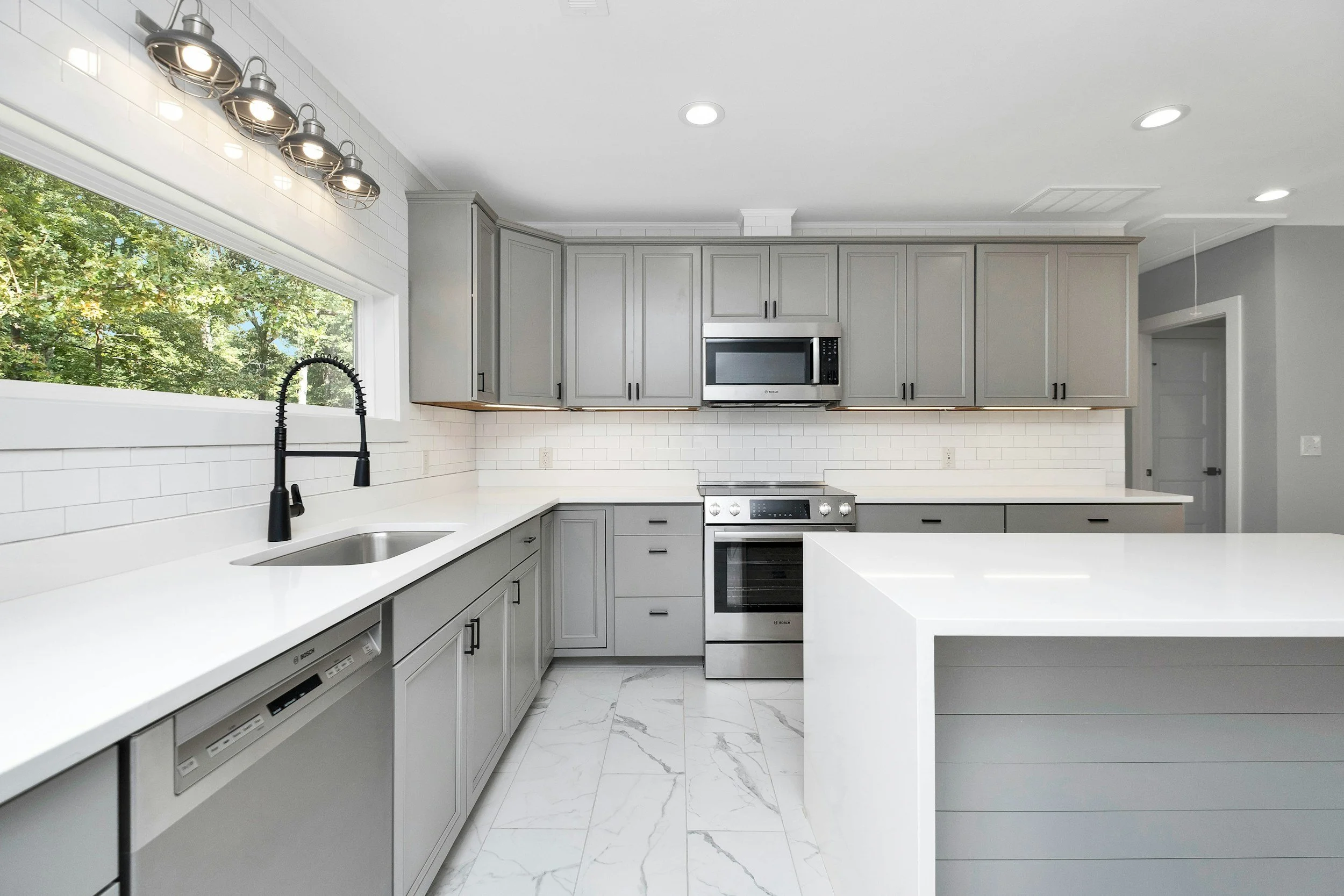 Modern kitchen with gray cabinets, white countertops, stainless steel appliances, a large window, and a white subway tile backsplash.