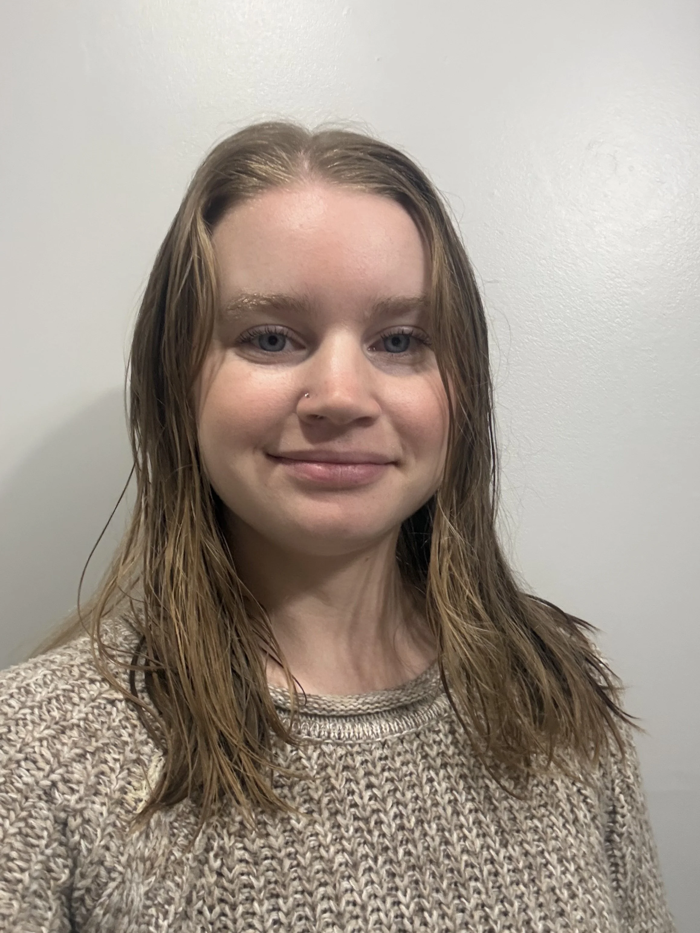 A young woman with light skin, blue eyes, and shoulder-length brown hair, smiling, wearing a beige knit sweater, and standing against a plain light-colored wall.