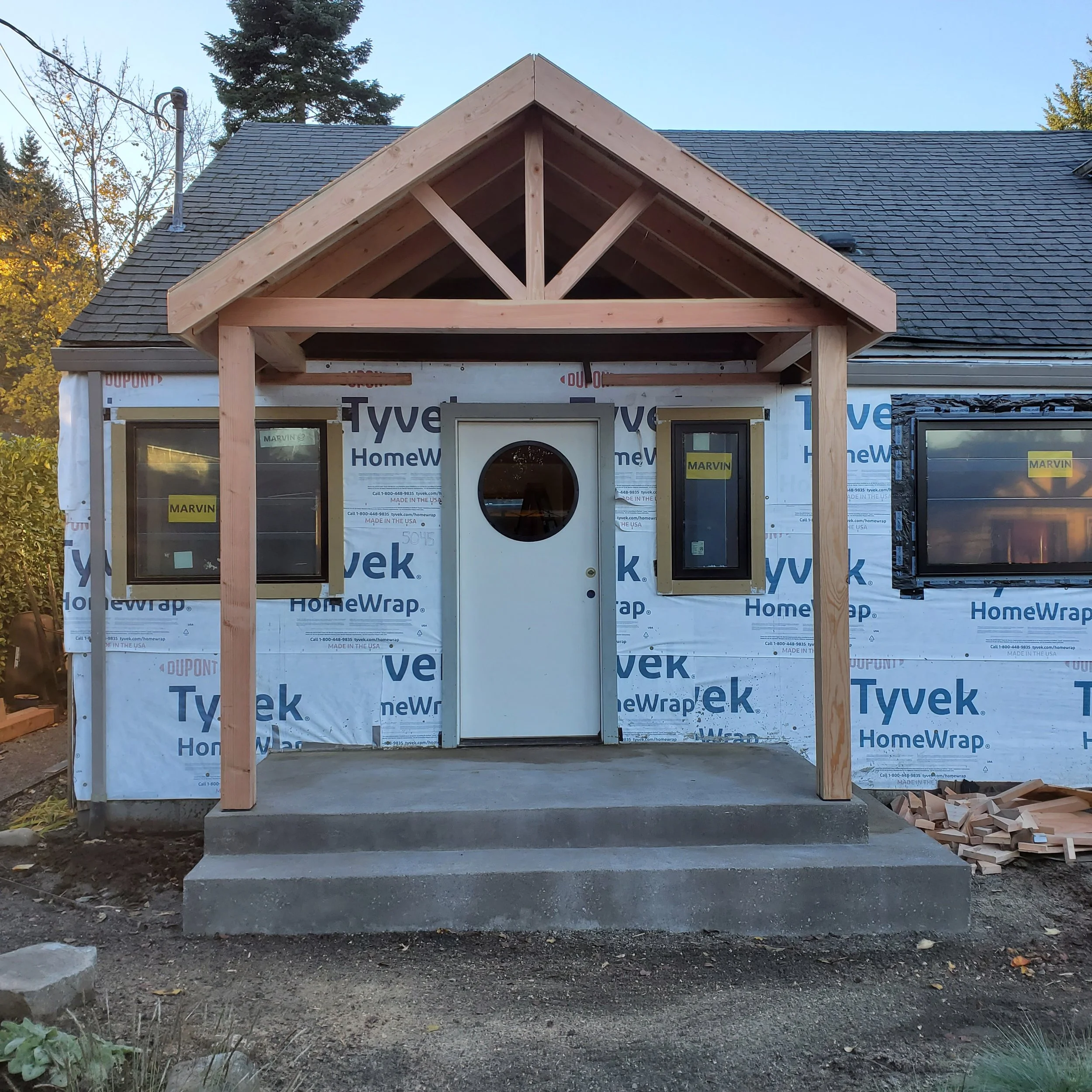 Front view of a house under construction with a small porch and a white door with a round window, surrounded by windows, with visible building materials and partial siding.