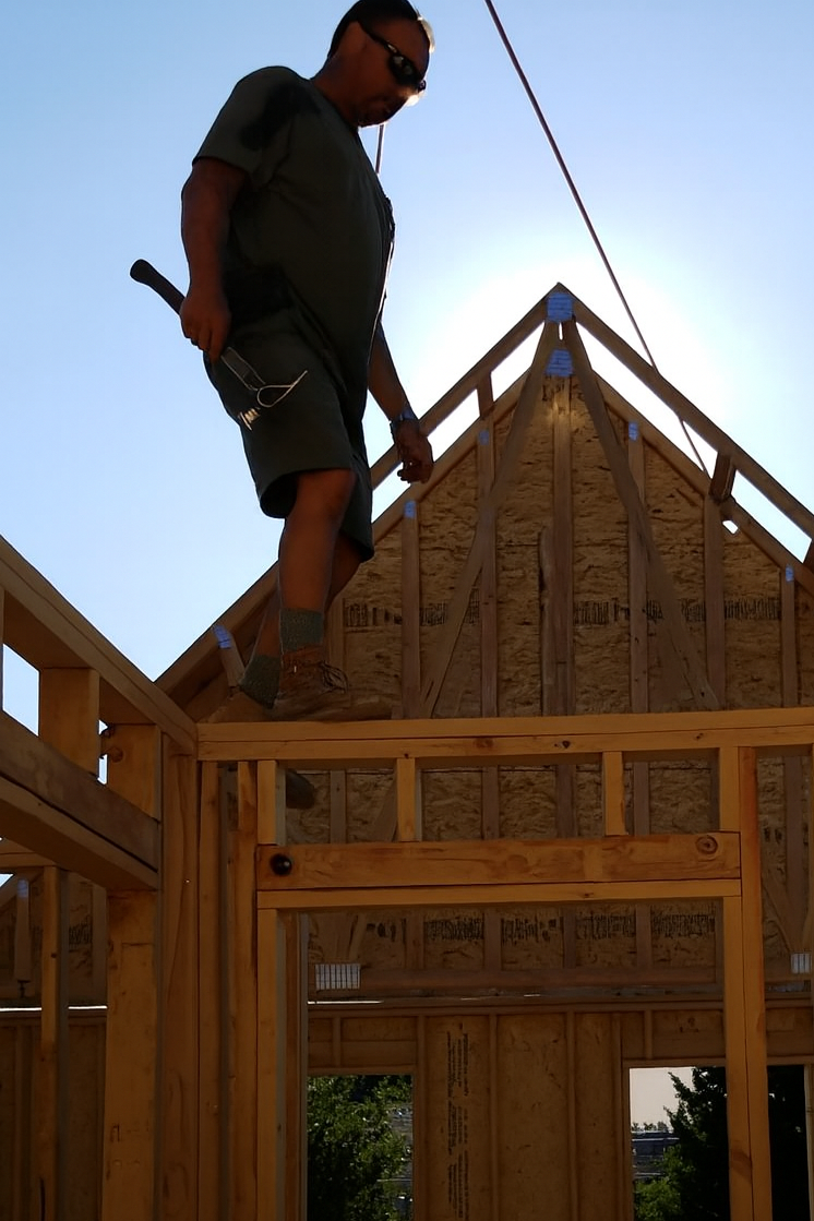 A man standing on the wooden frame of a house under construction, working on the roof structure during daytime.