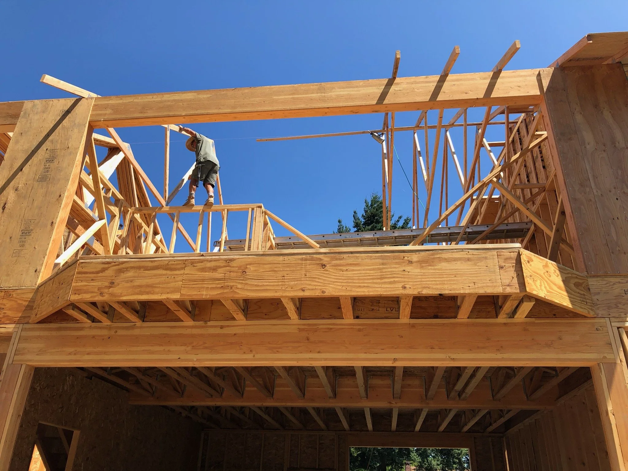 A construction worker standing on the wooden framing of a building under construction, using a tool to work on the upper part of the structure under a clear blue sky.