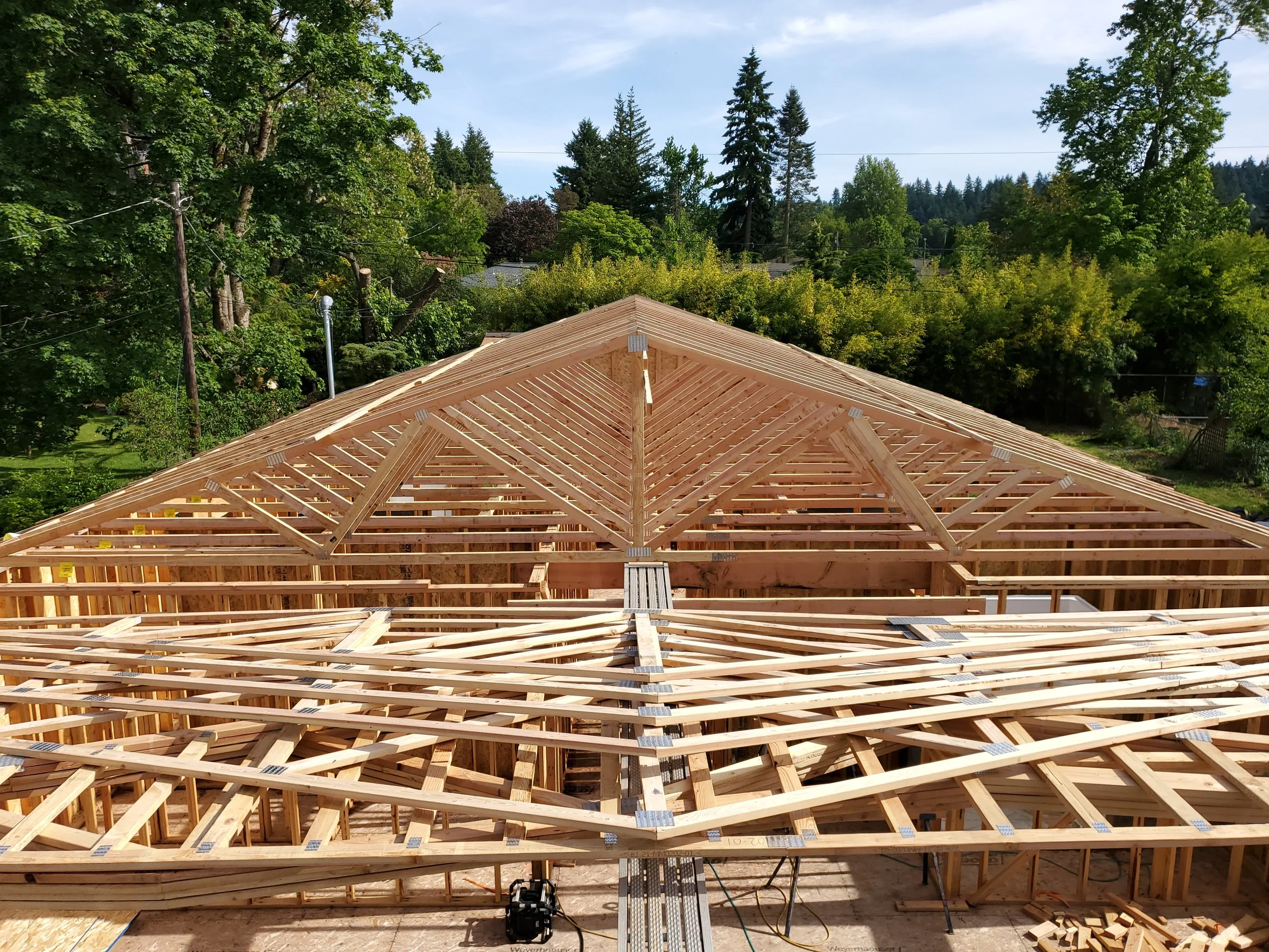 Construction of a wooden house frame in progress, with a view of the roof trusses and surrounding trees in the background.