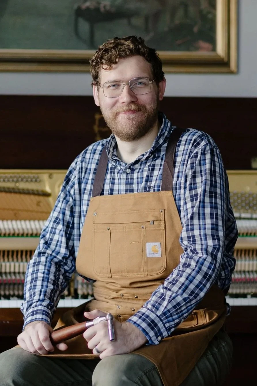Calvin W. Price smiling in a work apron, holding a tuning hammer, with an open piano in the background.