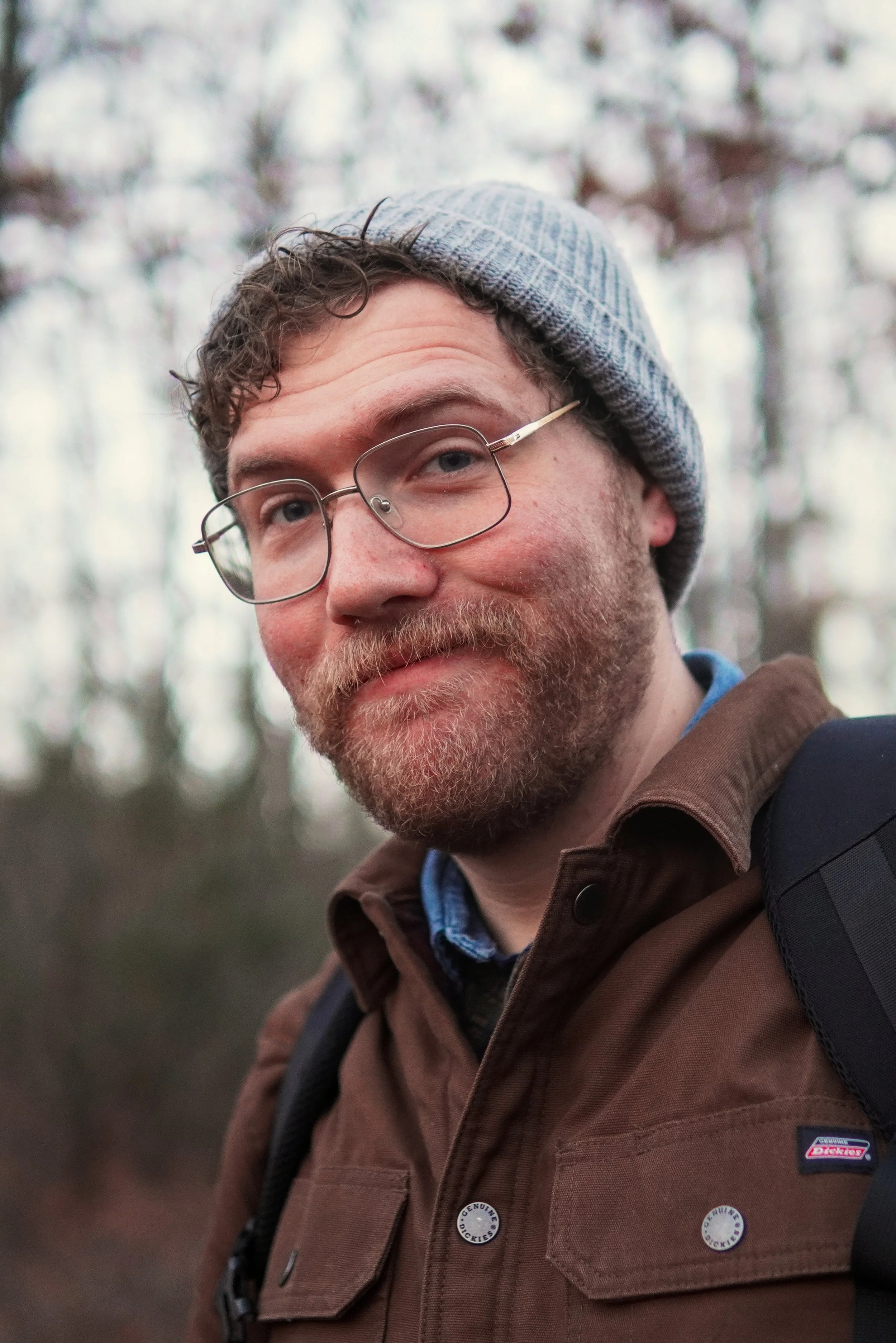 A young man with glasses and a beard, wearing a gray beanie and a brown jacket, looks at the camera with a slight smile while outdoors in a wooded area.