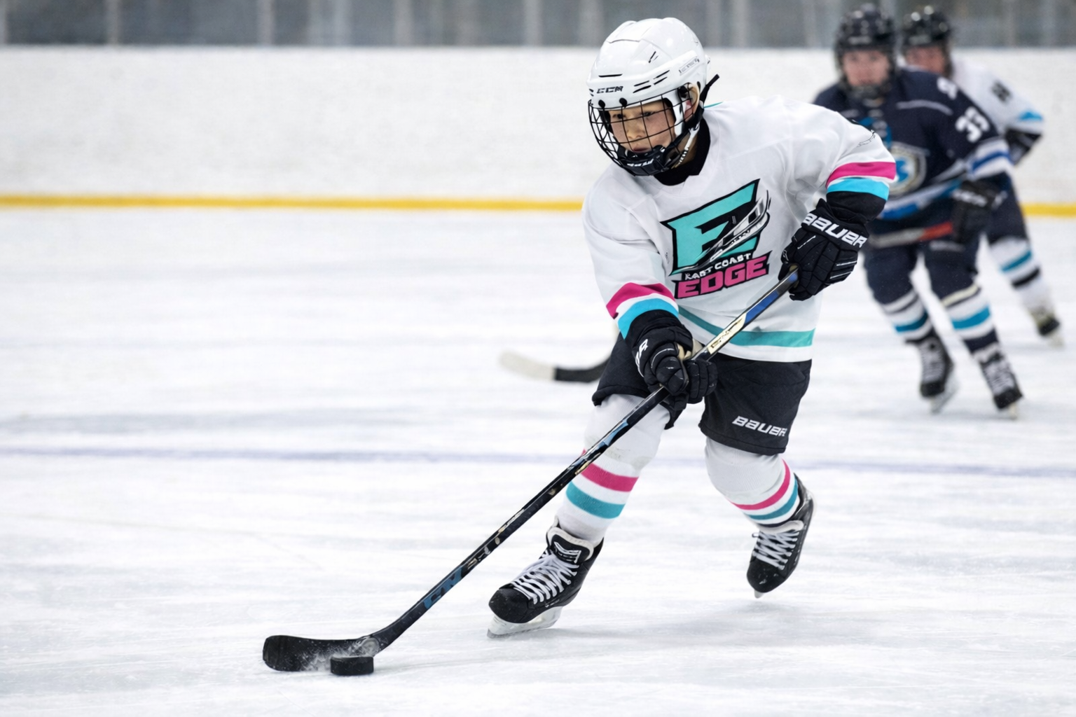 A young hockey player in a white jersey and helmet skates on the ice, controlling the puck with a hockey stick during a game.
