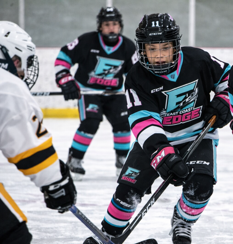 Youth ice hockey players face off during a game, wearing helmets, jerseys, and gloves, with one player in black and pink team gear, and the other in white and yellow.