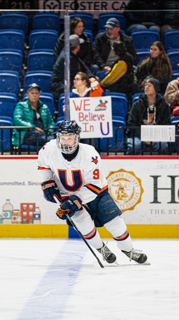 An ice hockey player in a white jersey with a large red and blue letter 'U' and the number 9 skating on the ice during a game. The player is wearing a helmet and gloves, holding a hockey stick, and appears focused. In the background, spectators are seated in blue bleachers, with some holding signs including one that reads 'WE Believe In U'.