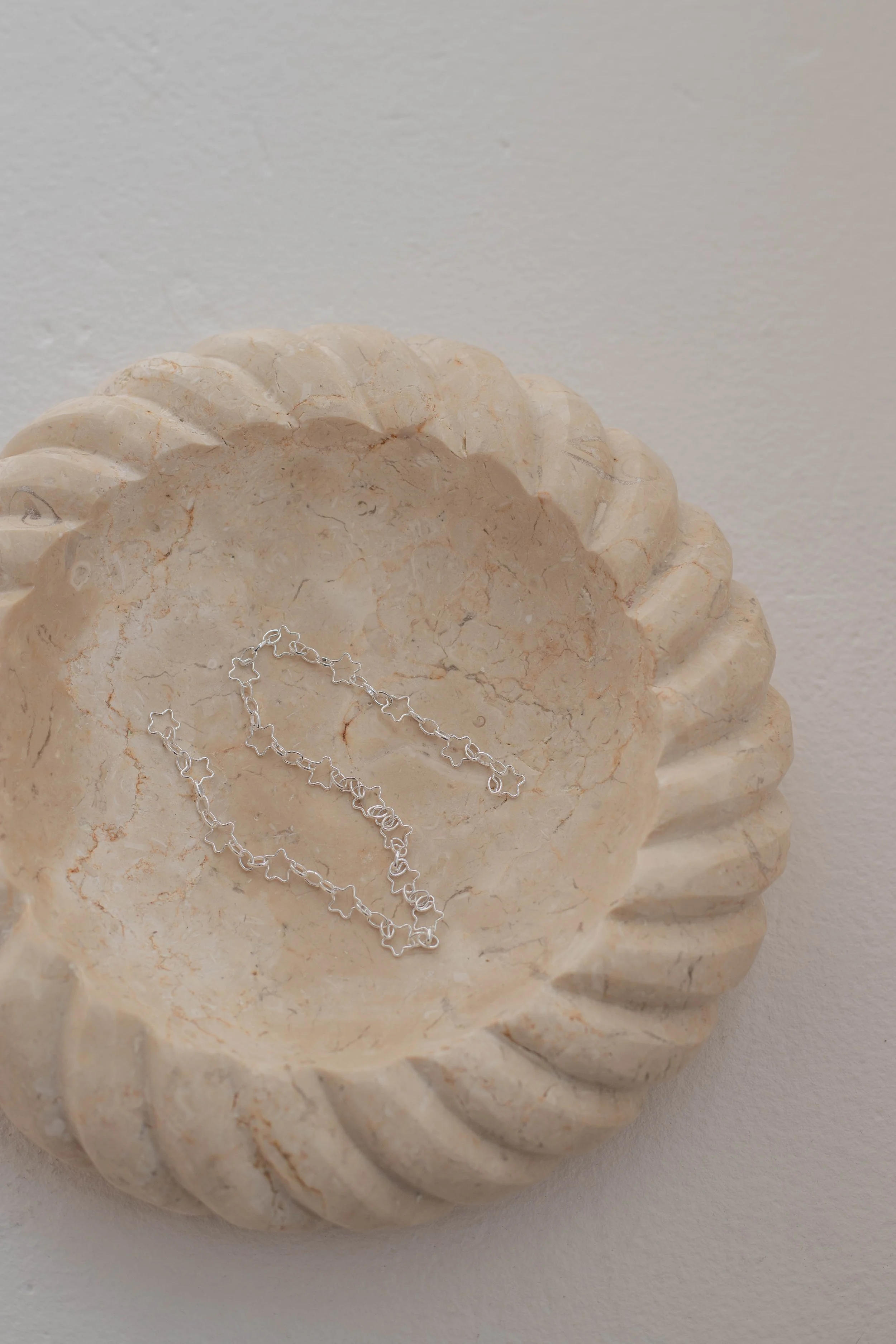 A marble shell-shaped dish with a silver star-shaped chain bracelet inside, placed against a plain off-white background.