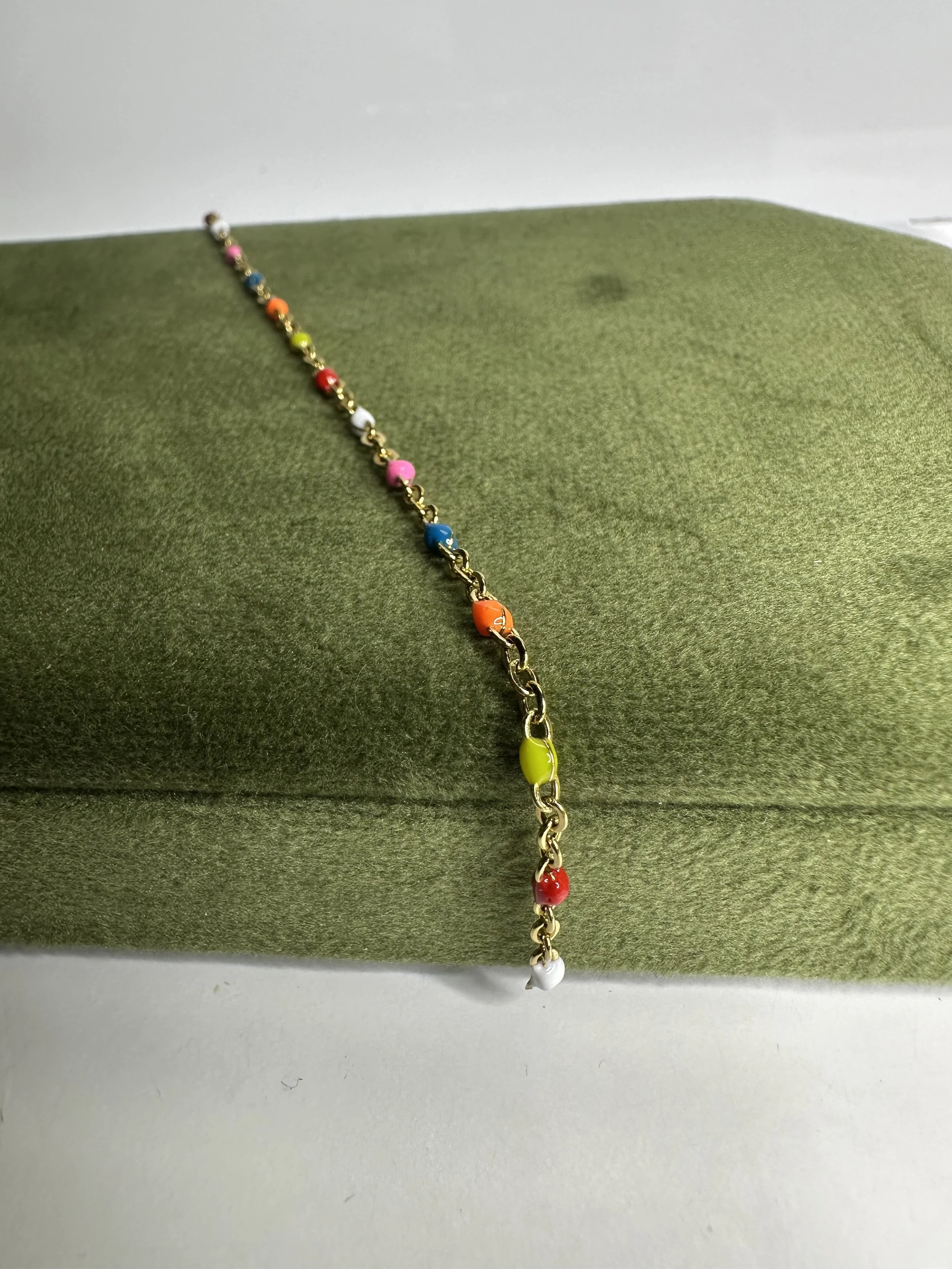 Colorful beaded bracelet with rainbow-colored beads on a gold chain, draped over a green fabric-covered box.