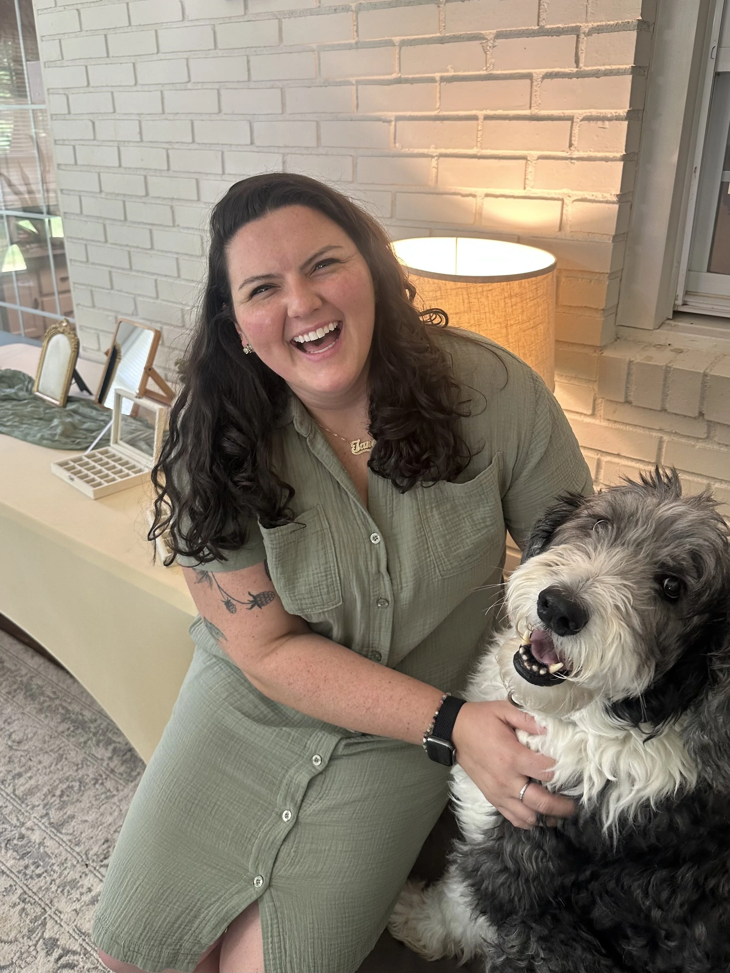 A woman with long dark curly hair, wearing a green dress with buttons, smiles happily while holding a black and white fluffy dog. She has a tattoo on her arm, wears a black watch, and a necklace. The background shows a white brick wall, a table with framed mirrors, and a lamp emitting warm light.